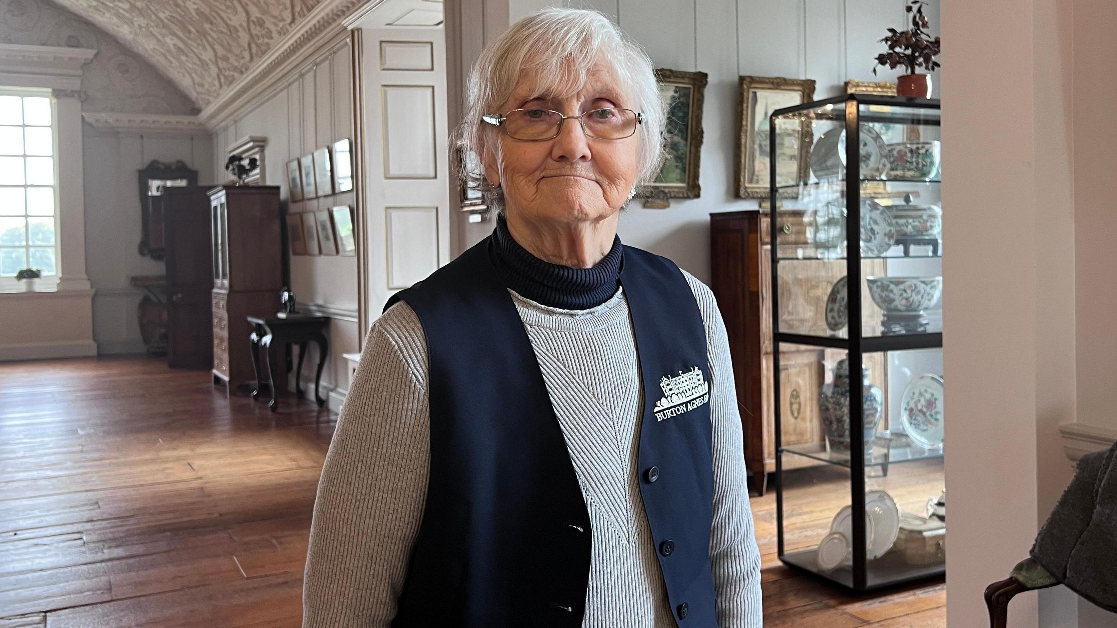 Head guide of Burton Agnes Hall, Pauline Waslin looks into the camera. She has short, white hair and silver-rimmed glasses. She is wearing a grey jumper with a navy-blue unbuttoned unic over top, it is branded with 'Burton Agnes Hall' with a small white drawing of the stately home.