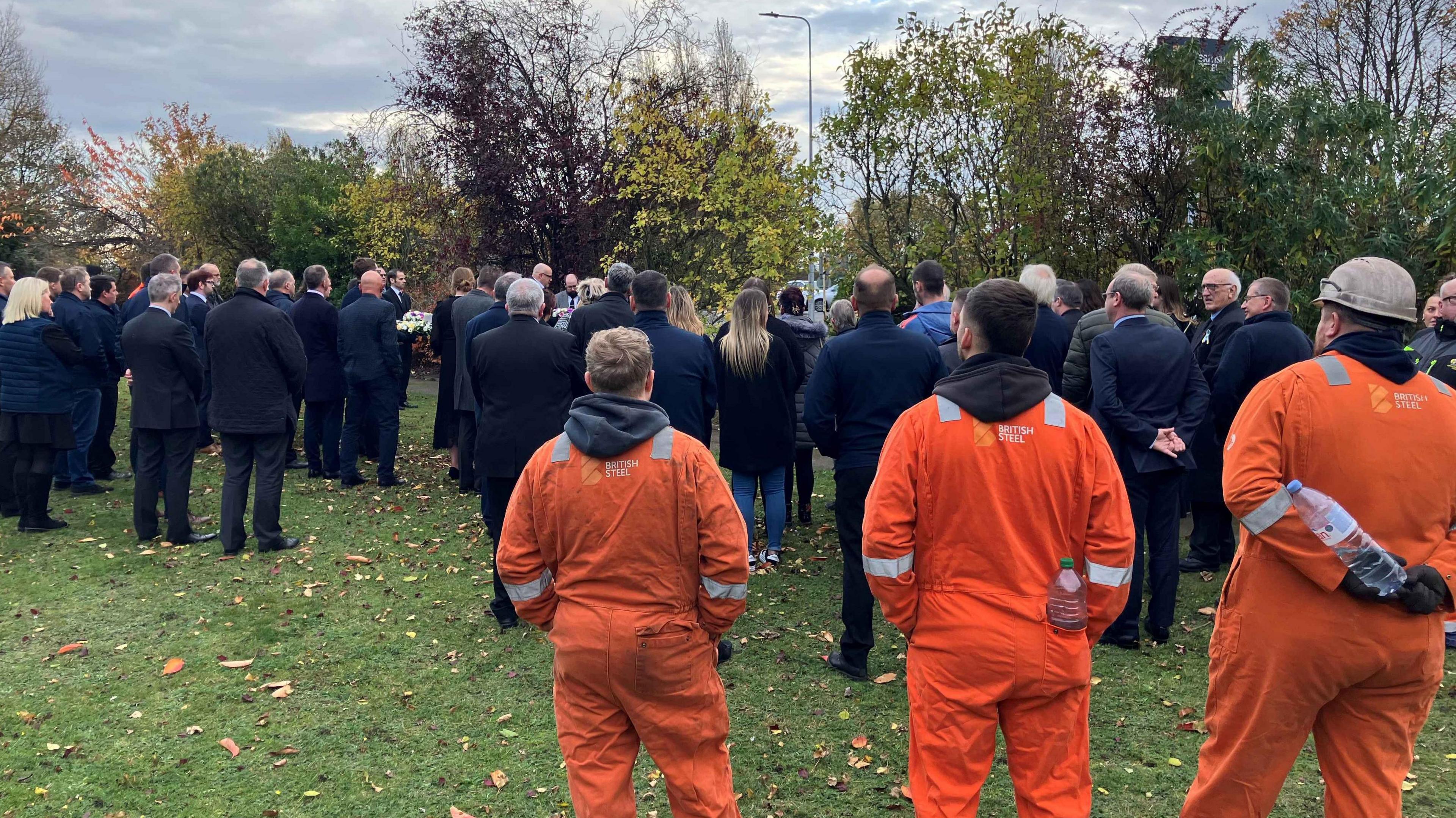 A rear view of people dressed smartly in dark colours standing on grass with three workers in orange overalls in the foreground watching a service.