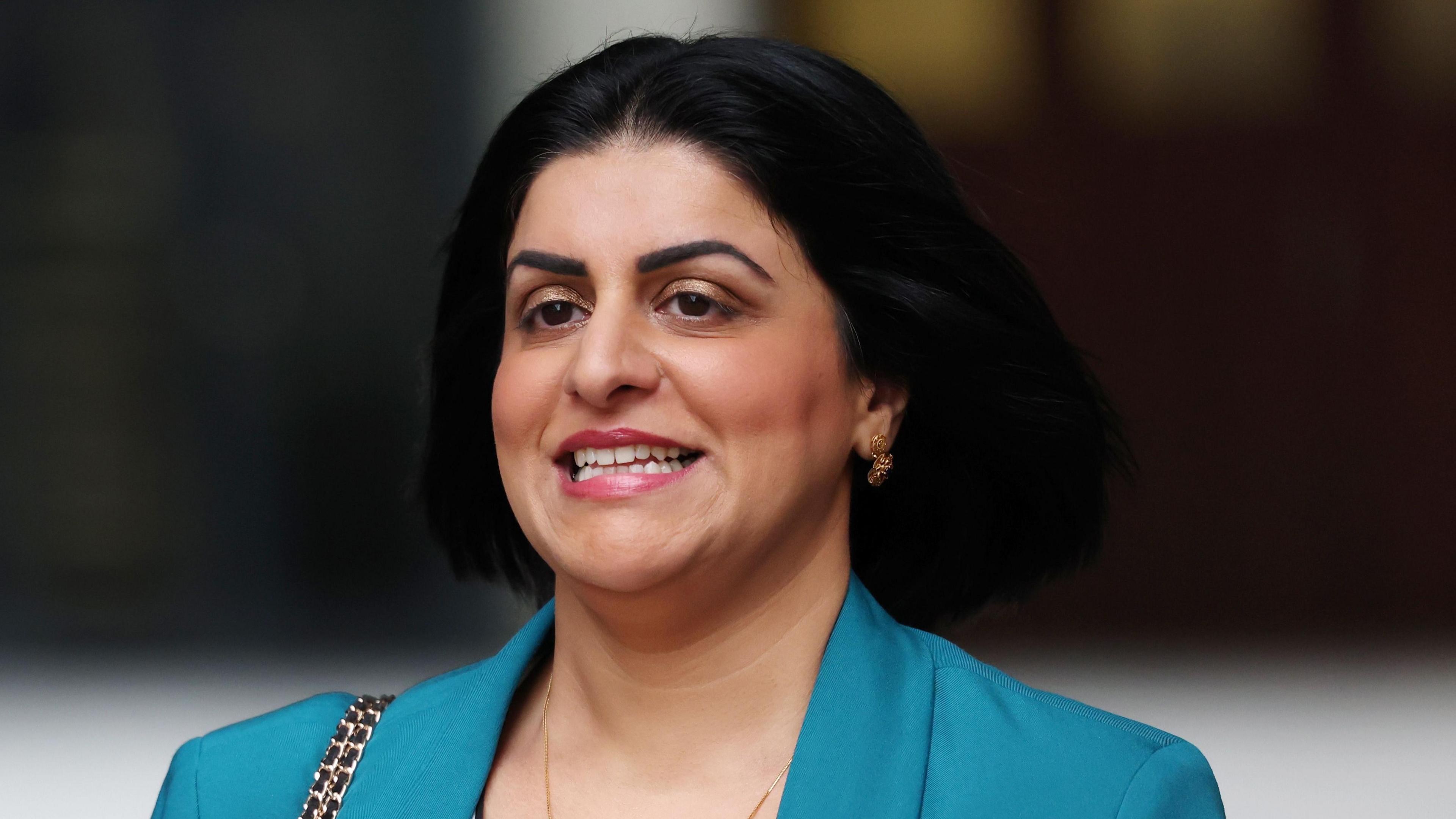 A woman with shoulder-length black hair is pictured wearing a bright blue blazer with short gold earrings