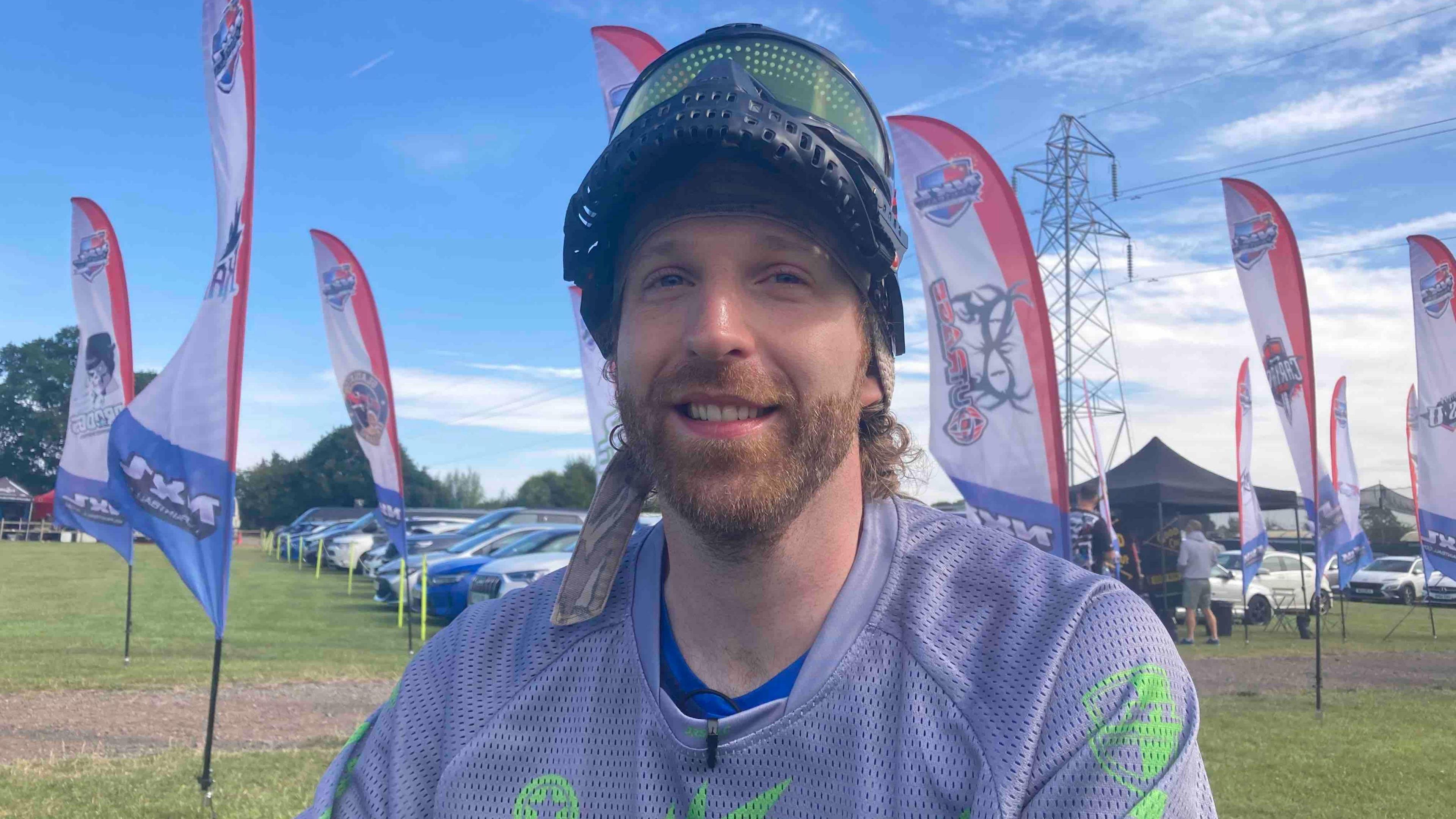 A man in a field. He has ginger hair and a ginger beard. He is wearing a black helmet and a grey sports top. There are flags and parked cars behind him.