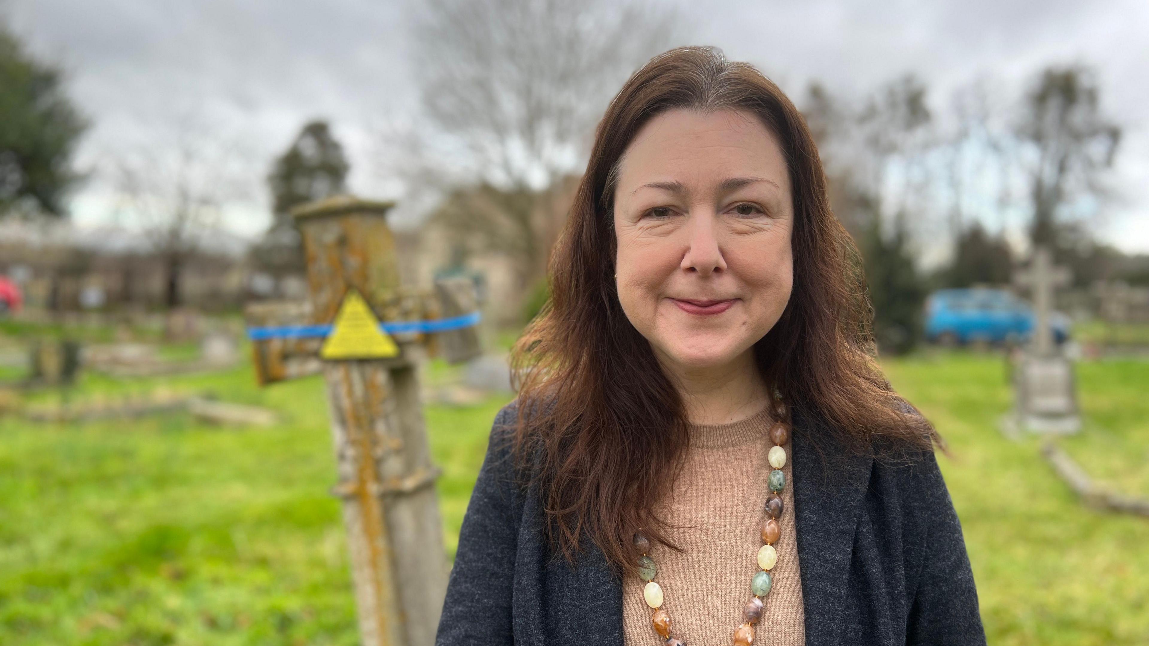 Stroud District Council leader, Chloe Turner, wearing a grey cardigan, brown top and brown, cream and green necklace, is standing in a churchyard.