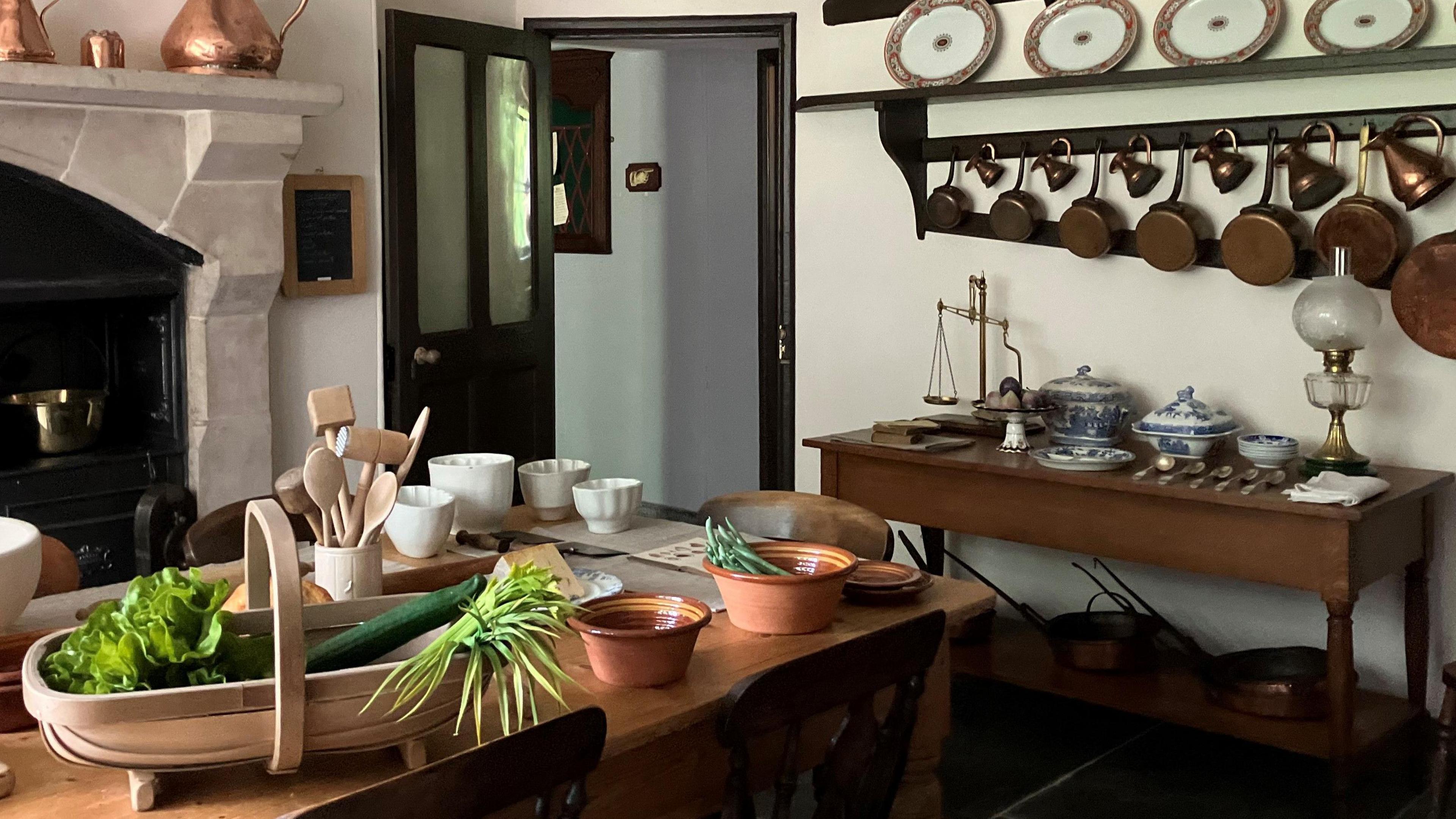 A renovated room in the rectory with pots and pans on the wall, a table with  crockery and a vegetable basket and a fireplace and door in the background. 