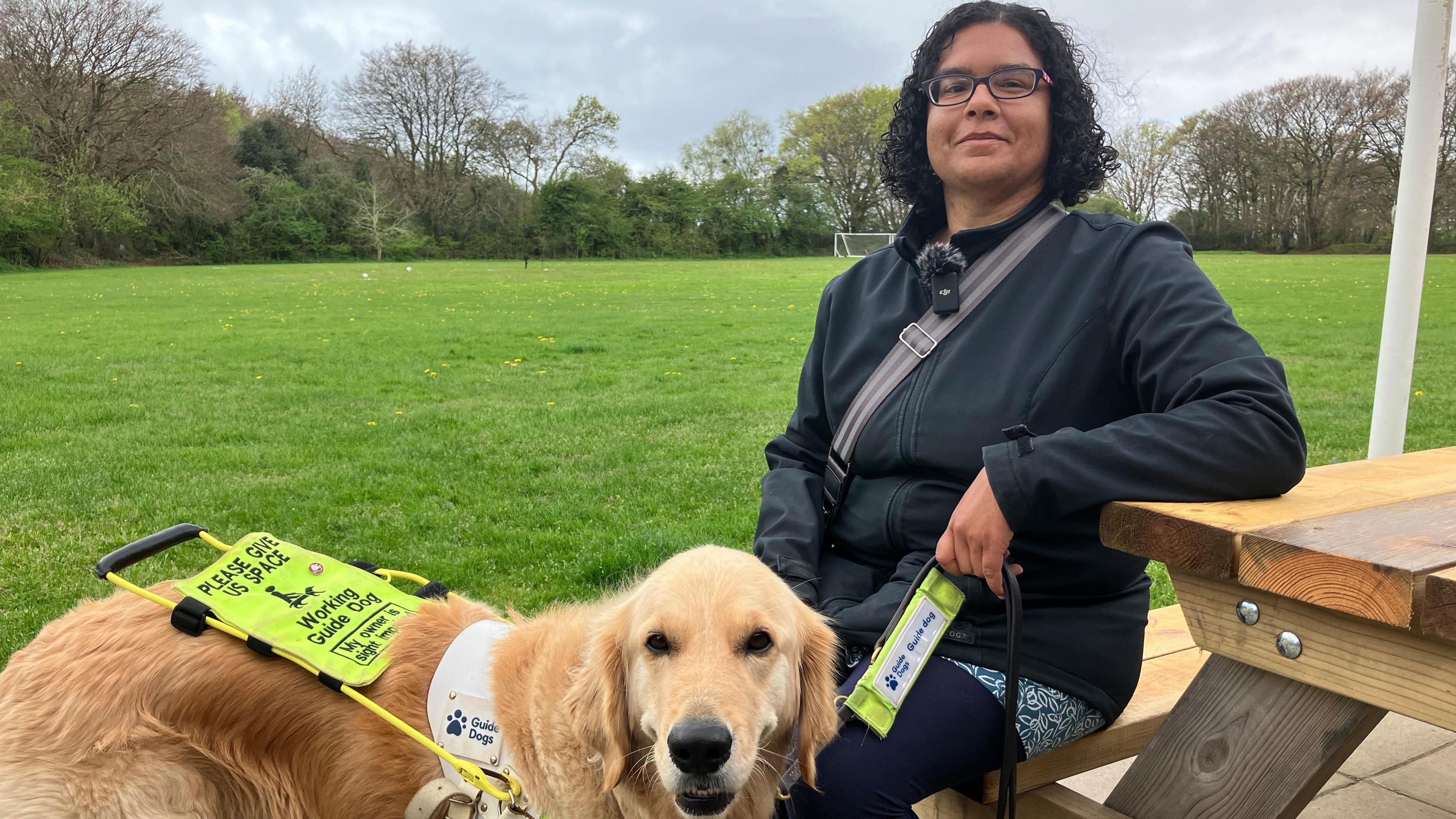 A woman in a black jacket sitting on a picnic bench in front of a field with a golden Labrador standing in front of her. The dog is wearing a harness describing it as a a working guide dog.