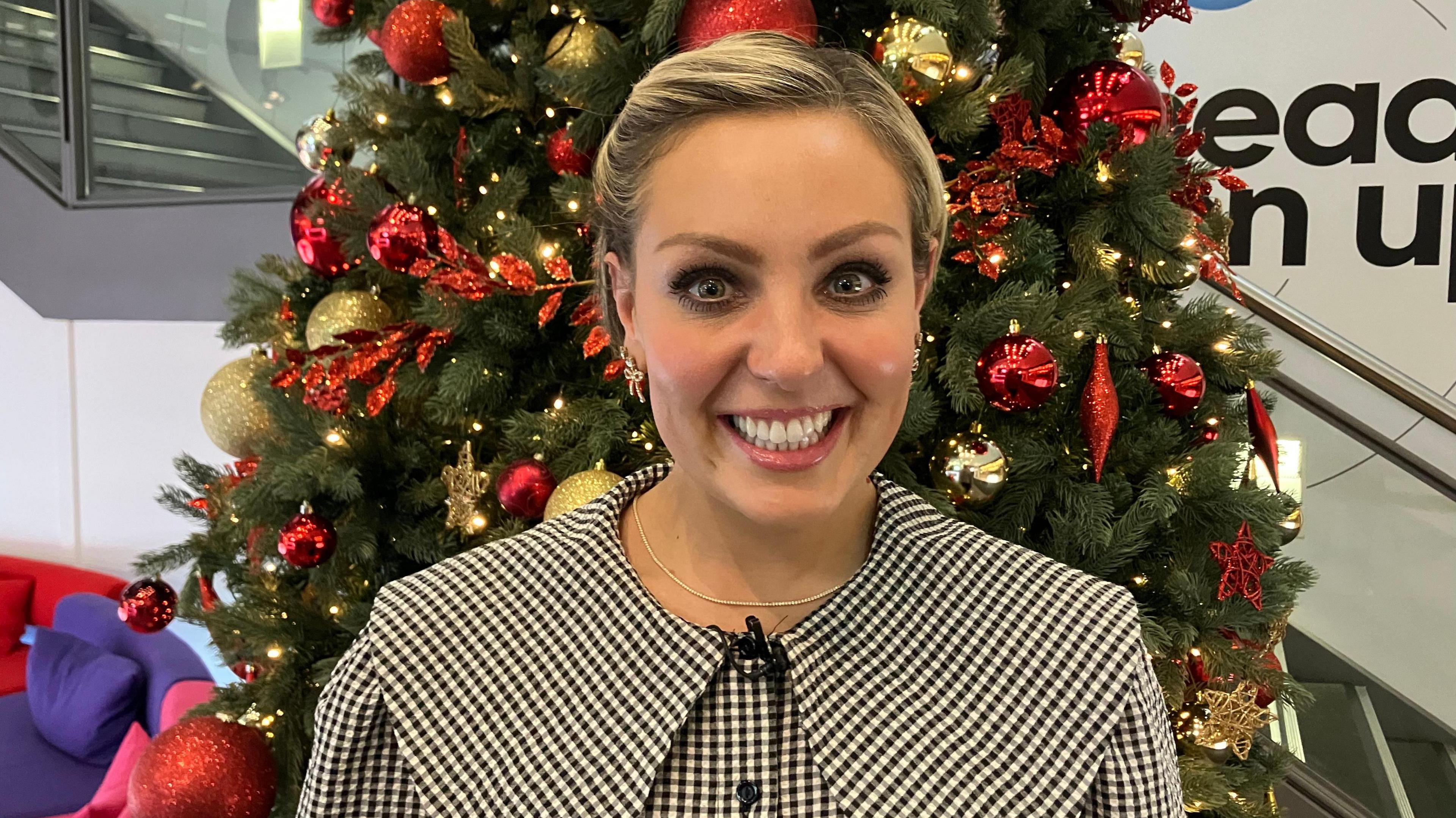 Amy Dowden smiling looking into the camera. She is wearing chequered dress and a gold necklace is also visible. Behind her, a Christmas tree is decorated with baubles and festive lights.