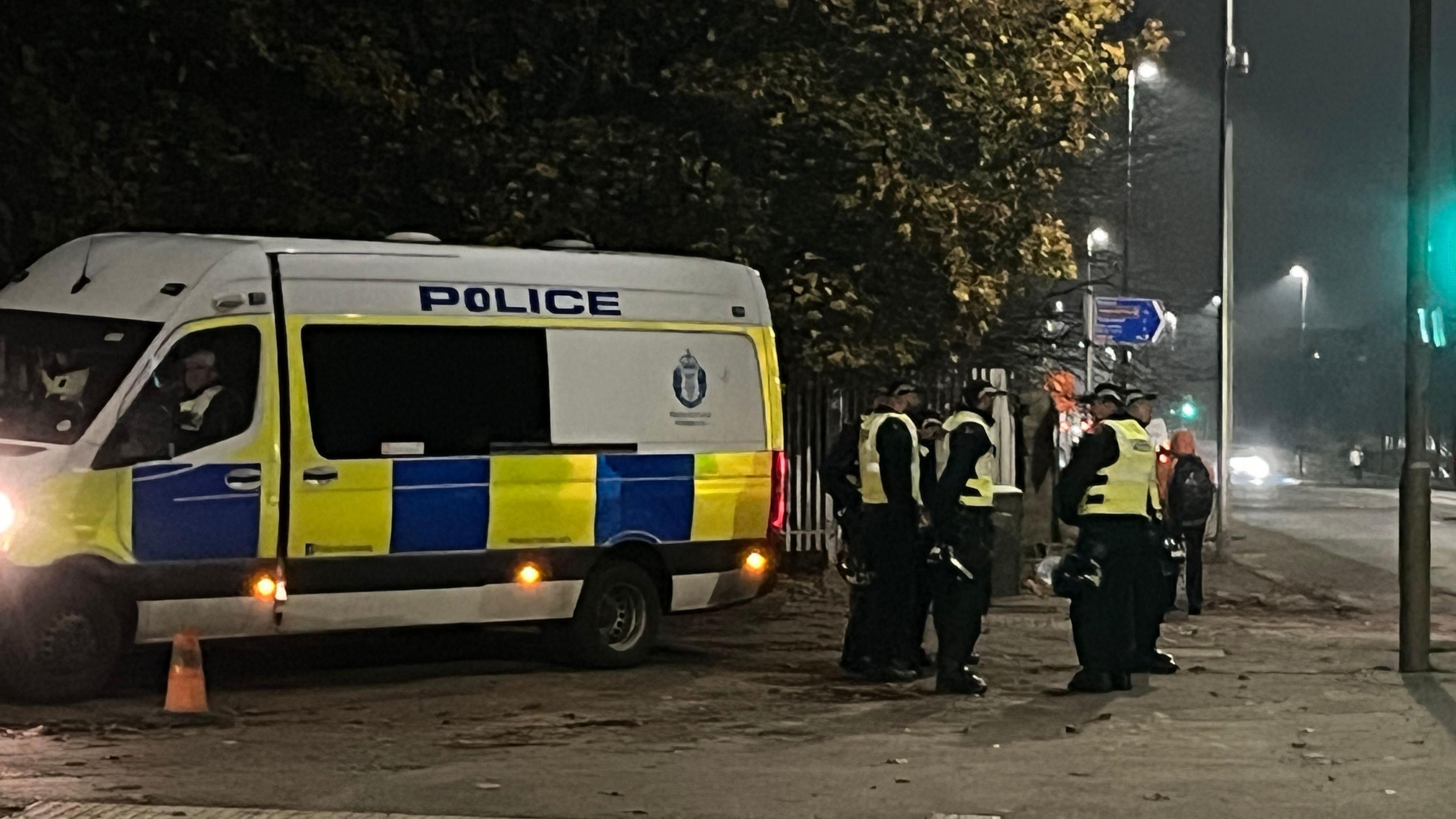 A police van parked on a kerb at night with four officers standing nearby holding helmets. 