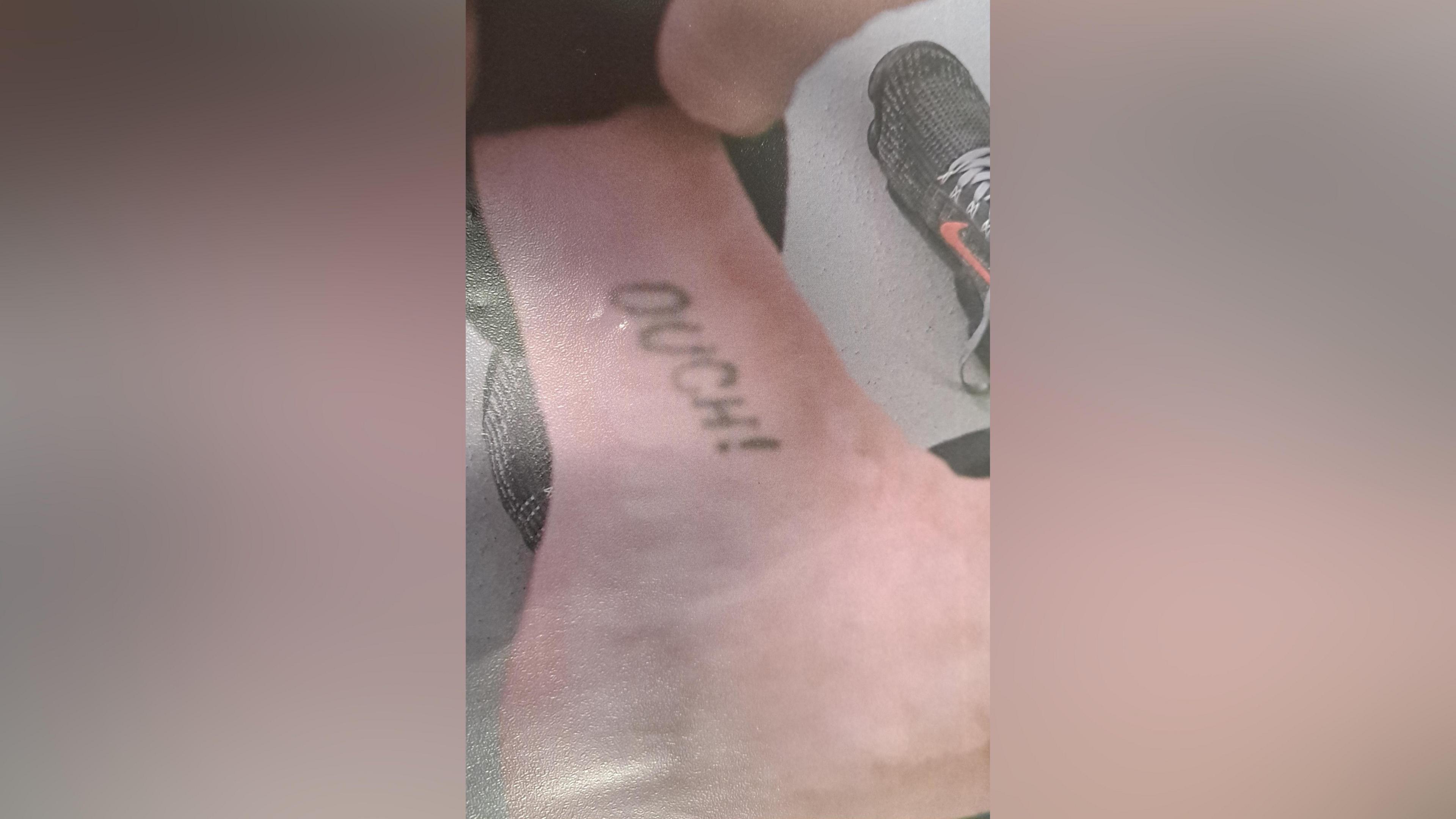 Close up of the word "ouch!" on the ankle of a man leading on to his foot. A black trainer with a red Nike tick can be seen beyond.