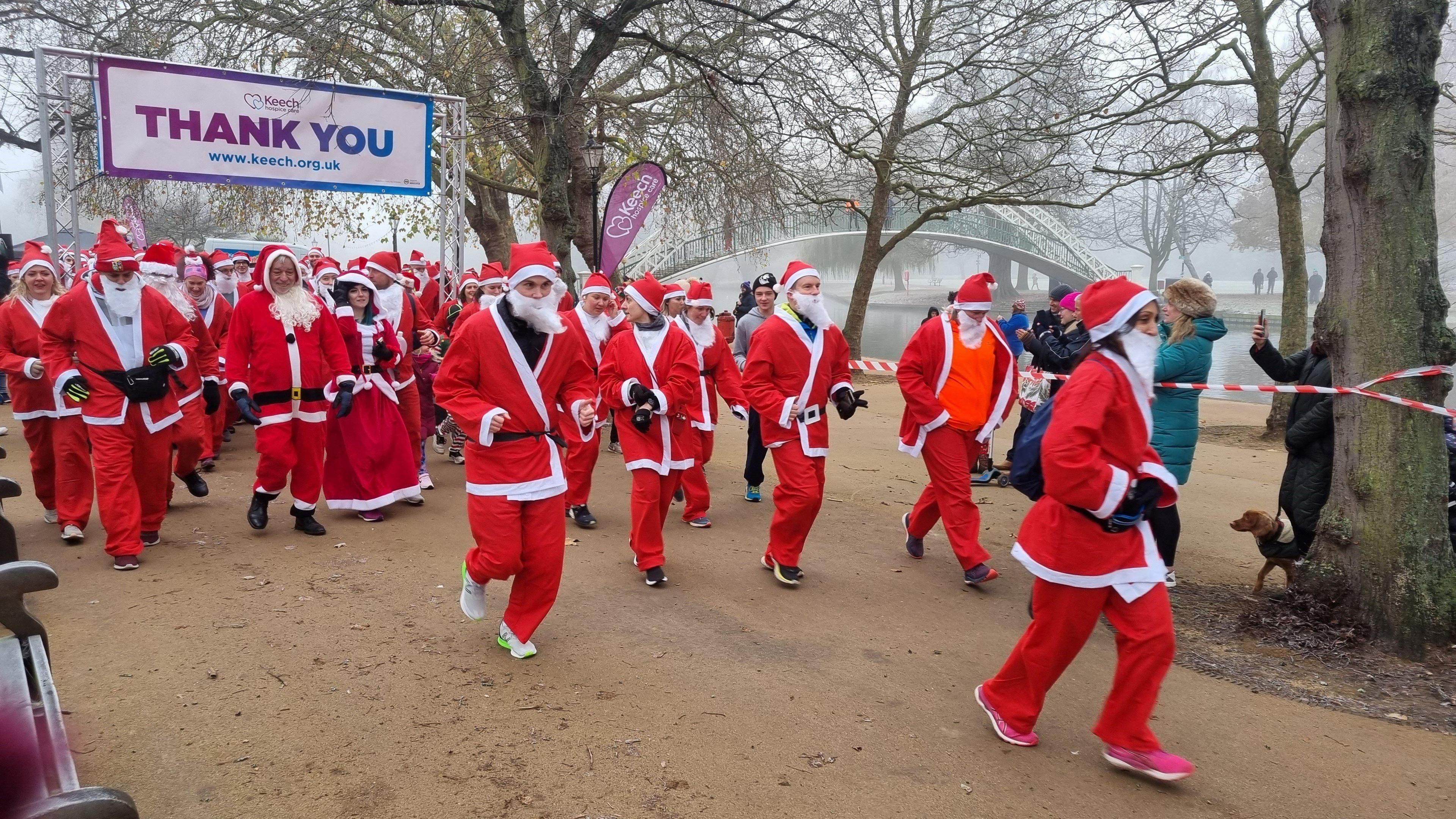 A large number of people running in a Santa Run, wearing red and white Santa suits with hats and trainers on. Spectators are taking photos, and there are dogs, too. A river is in the distance, along with a white suspension bridge. The sky is misty and there trees, with no leaves on, along the river