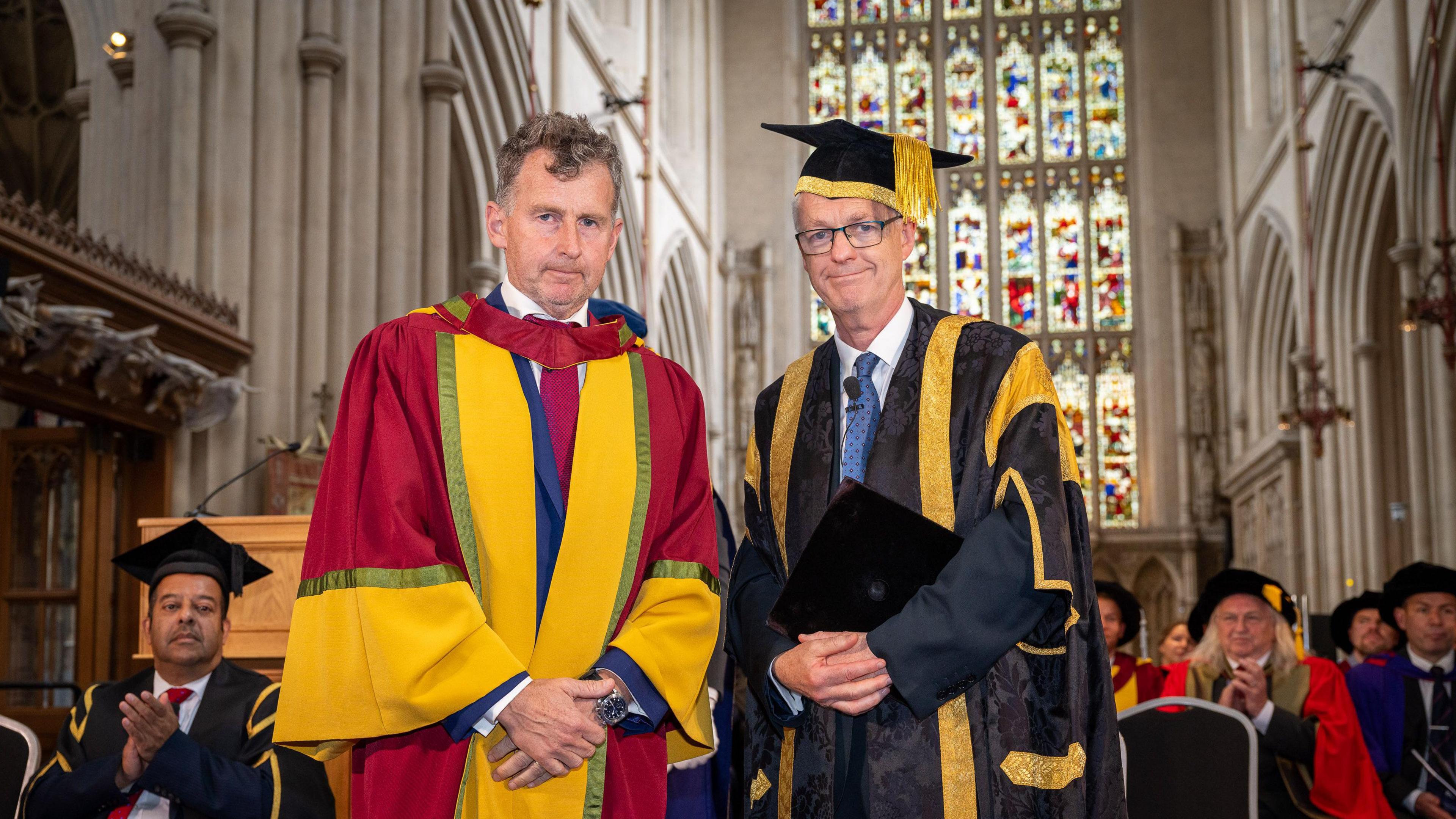 Two men wearing red and golden graduation robes, smiling with their hands closed in front of them, with standing in large hall with stained glass windows and other university officials behind them.