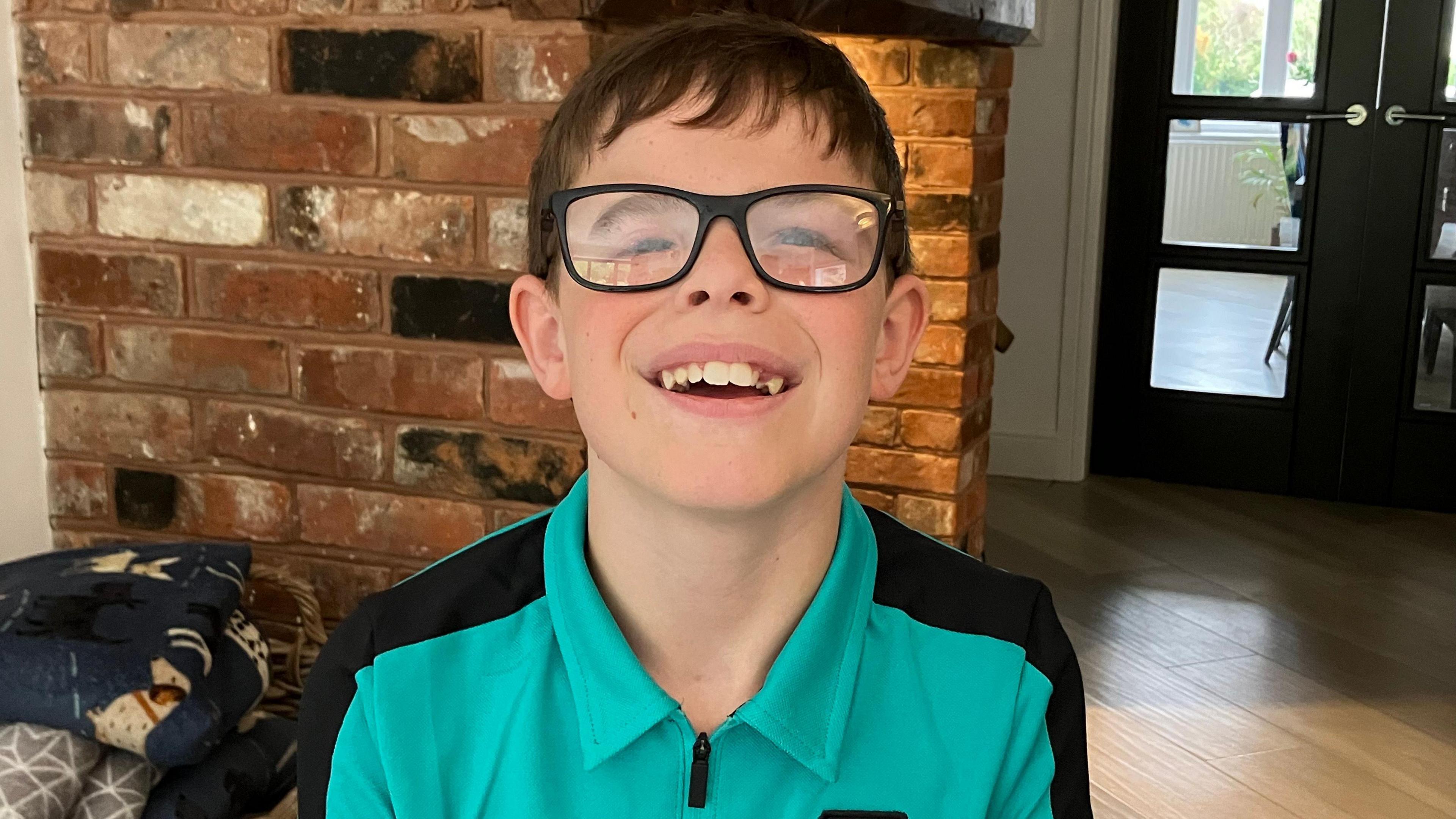 A boy with short black hair, glasses, a green and black shirt, smiles as he stands in a house. A brick wall is behind him as well as a door leading into another room.
