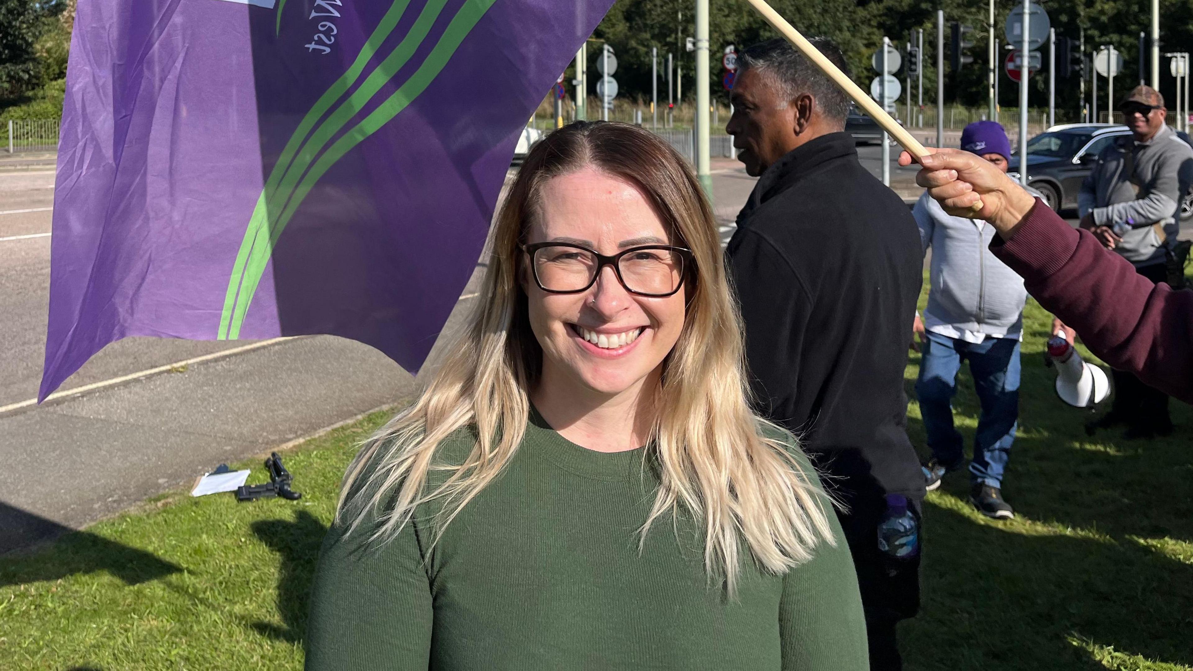 Jayne Jackson who is smiling at the camera. She has on black rimmed glasses and a green top. Behind her are people holding purple and green Unison flags. 