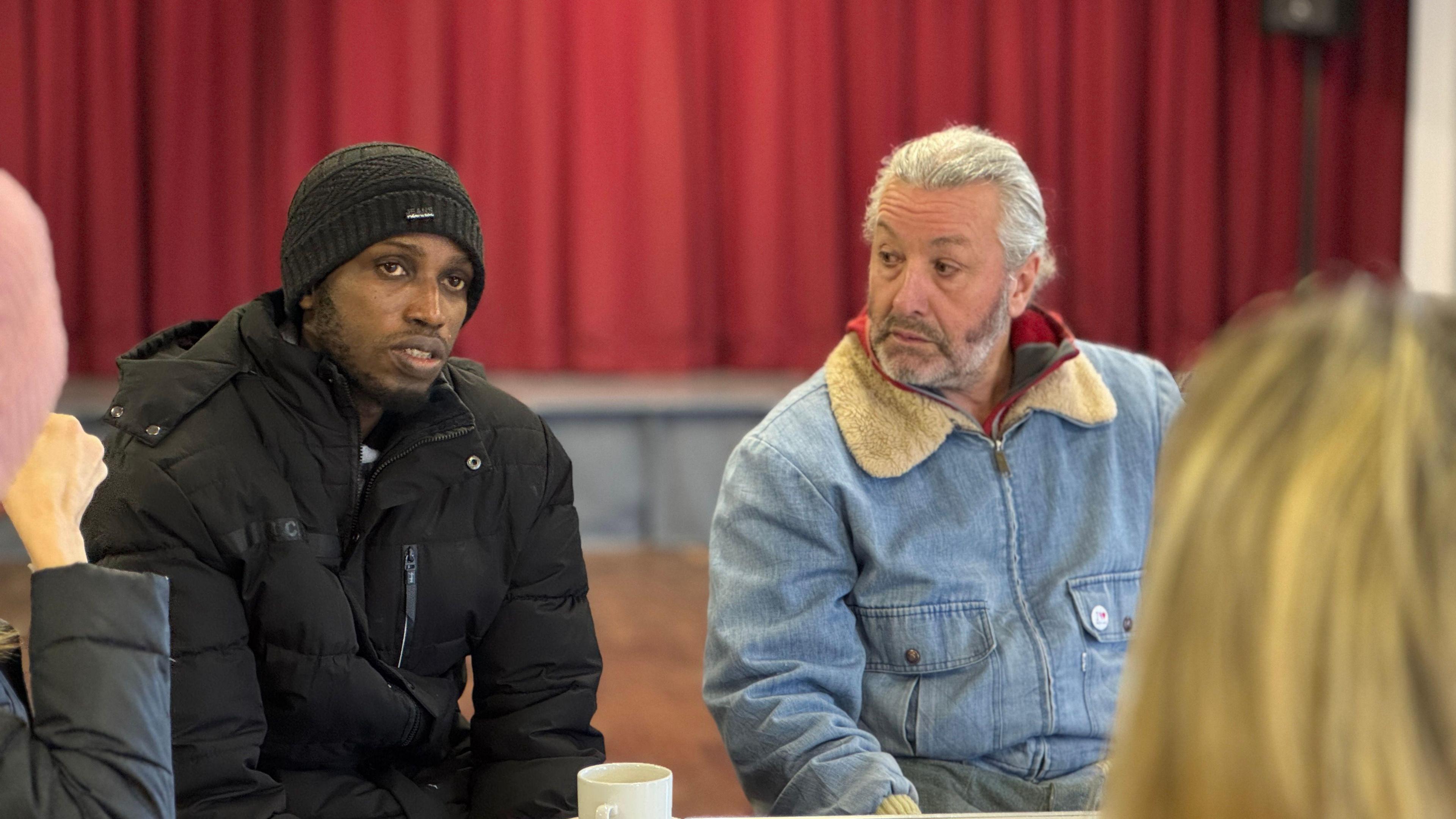 Two men are sitting down. The man on the left is wearing a black beanie and a black winter coat. The man on the right is look at the other man, he is wearing a faded blue denim coat with a white woolen collar. Behind them is what appears to be a stage with a large red curtain