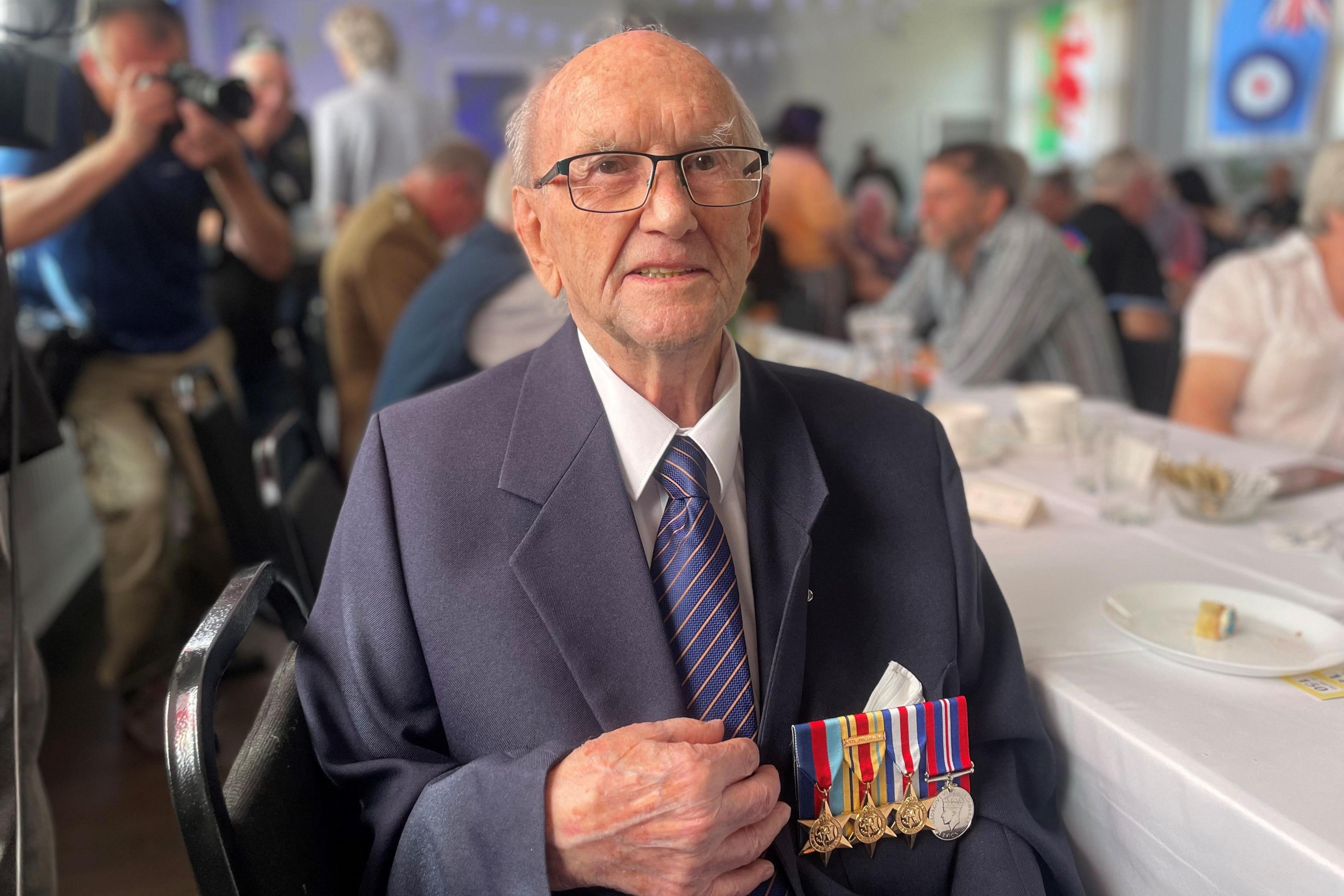 Mr Bird sat on a chair, there are people sat on tables around him, as well as photographers. He is wearing a blue suit, with a white shirt and blue and red striped tie. His new medals are pinned on the left side of his chest.