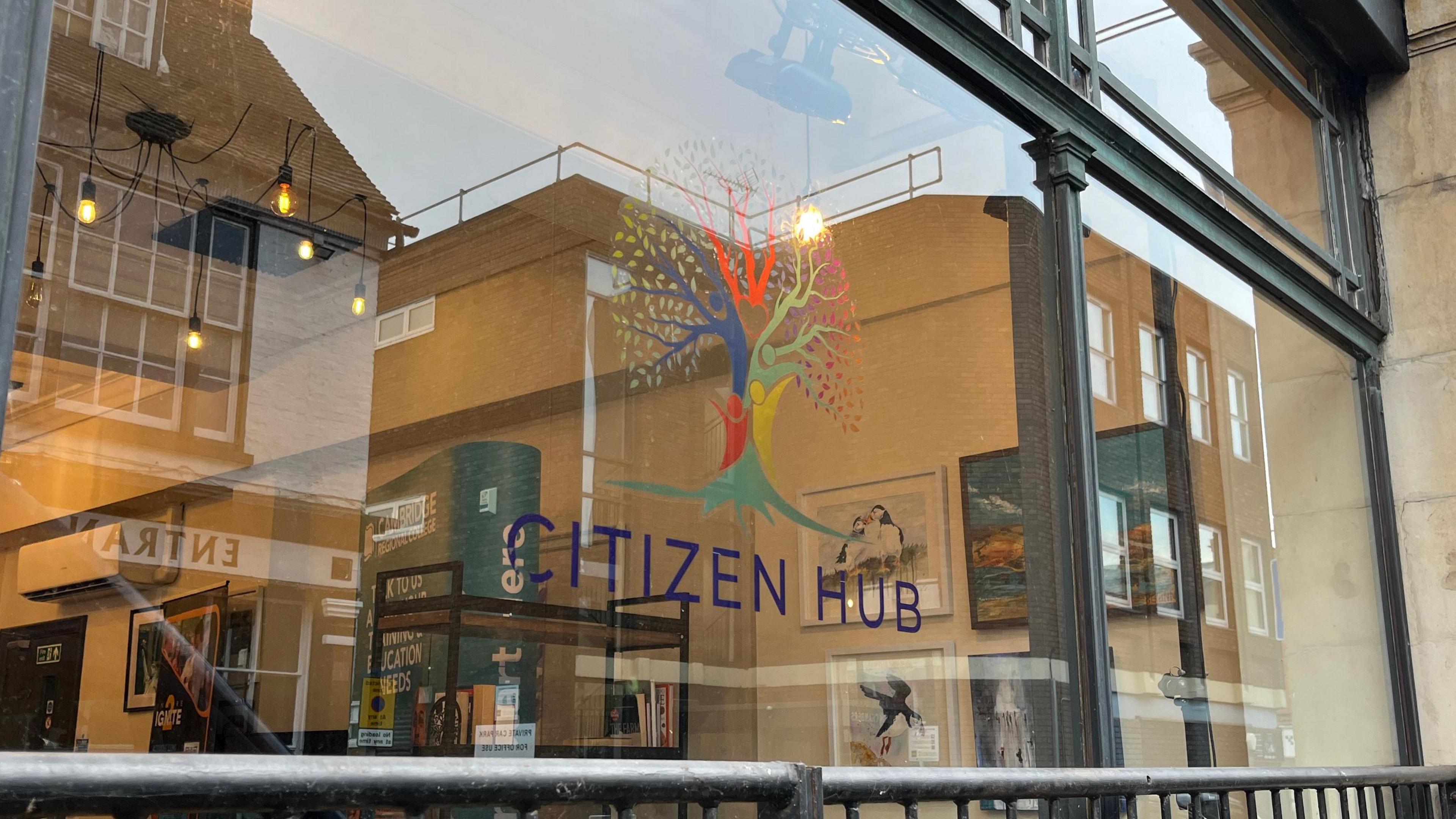 A window looking into the Citizen Hub. There is a logo and the words Citizen Hub printed on the glass. Inside there are photos of the wall, paintings and bookshelves. 