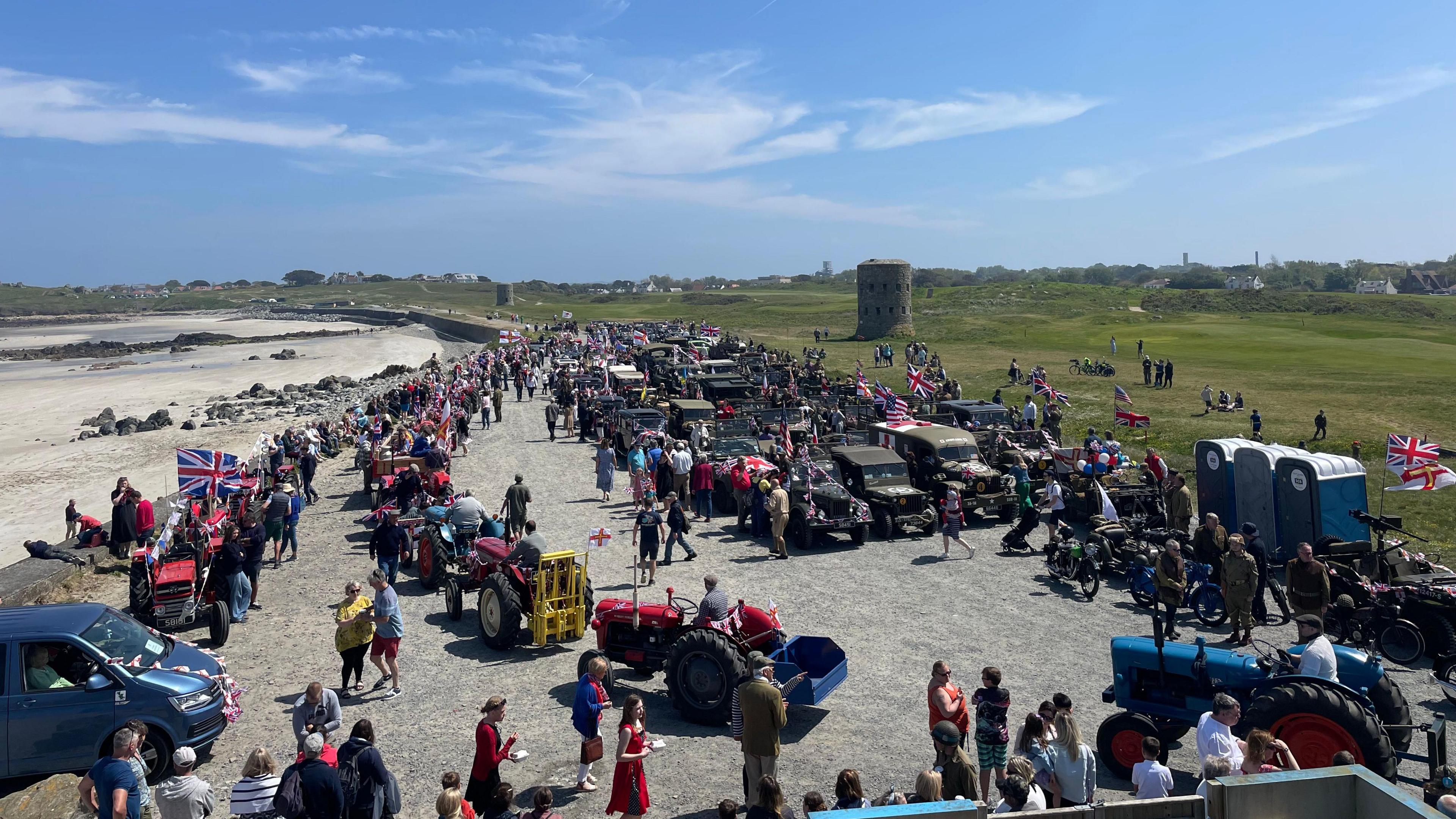 Vehicles and people park up for a break near the seafront during last year's cavalcade in Guernsey Tractors and 4X4's took part. People are holding Union Jack flags. A large grass area is behind the seafront.