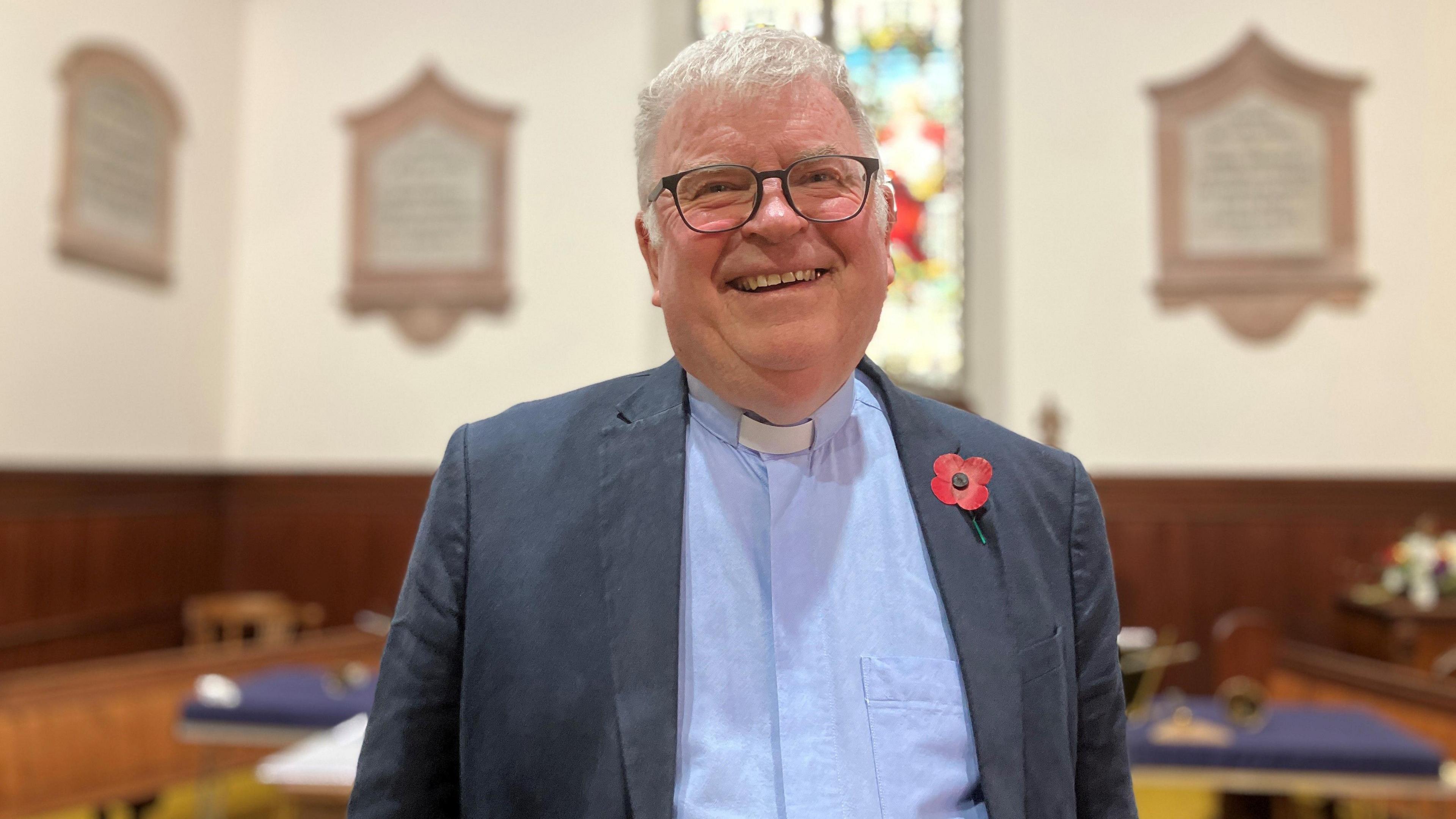 A church of Scotland minister stands inside a church. He has grey hair and glasses and is wearing a blue shirt with a dog collar and a jacket with a poppy on it.