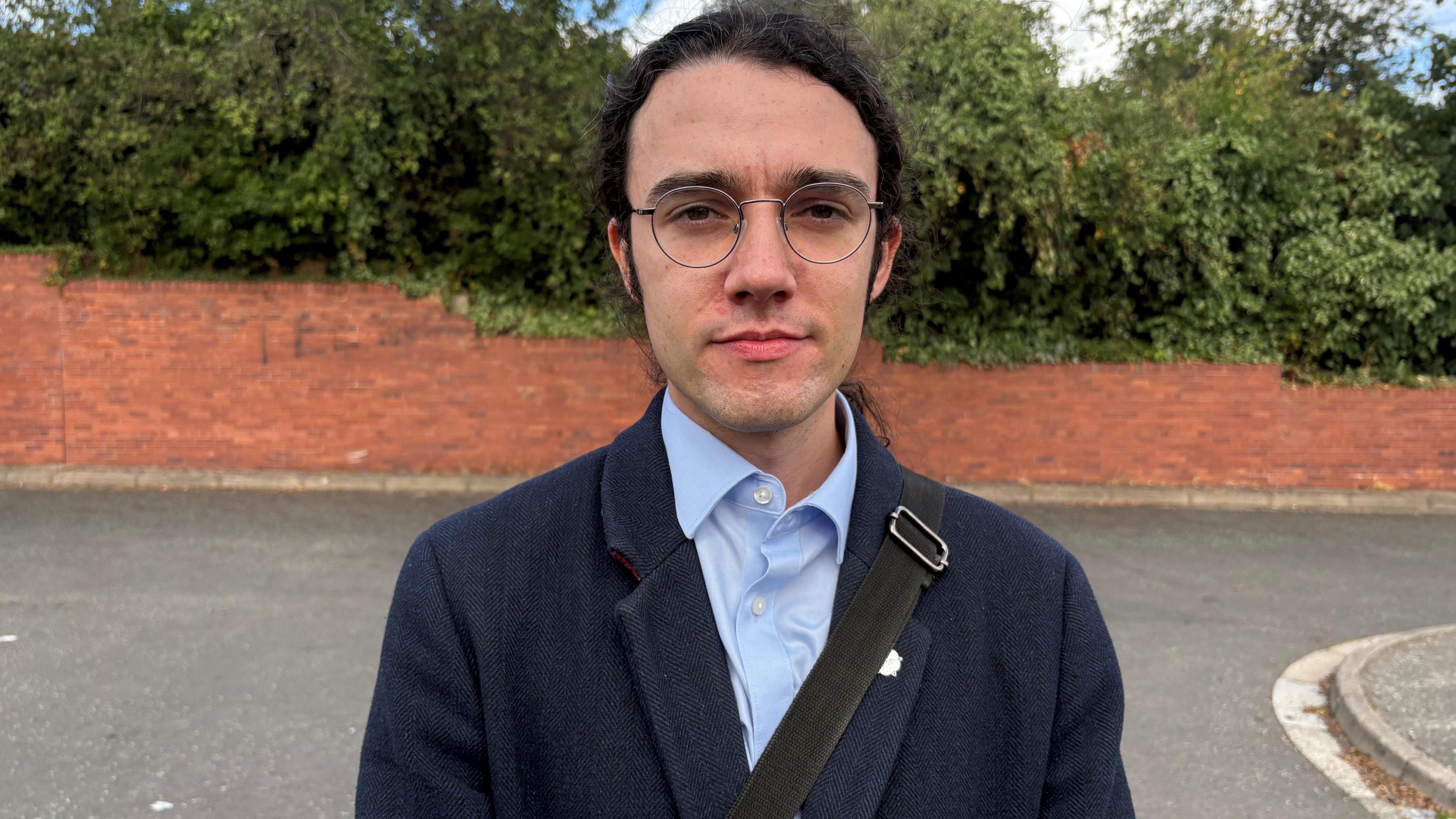 A man with long black hair, tied back, is wearing circular-rimmed glasses, a light blue shirt, and a dark blue jacket, with a strap of a bag going across his body. He is standing on a street. Behind him is a brick wall with trees behind it.