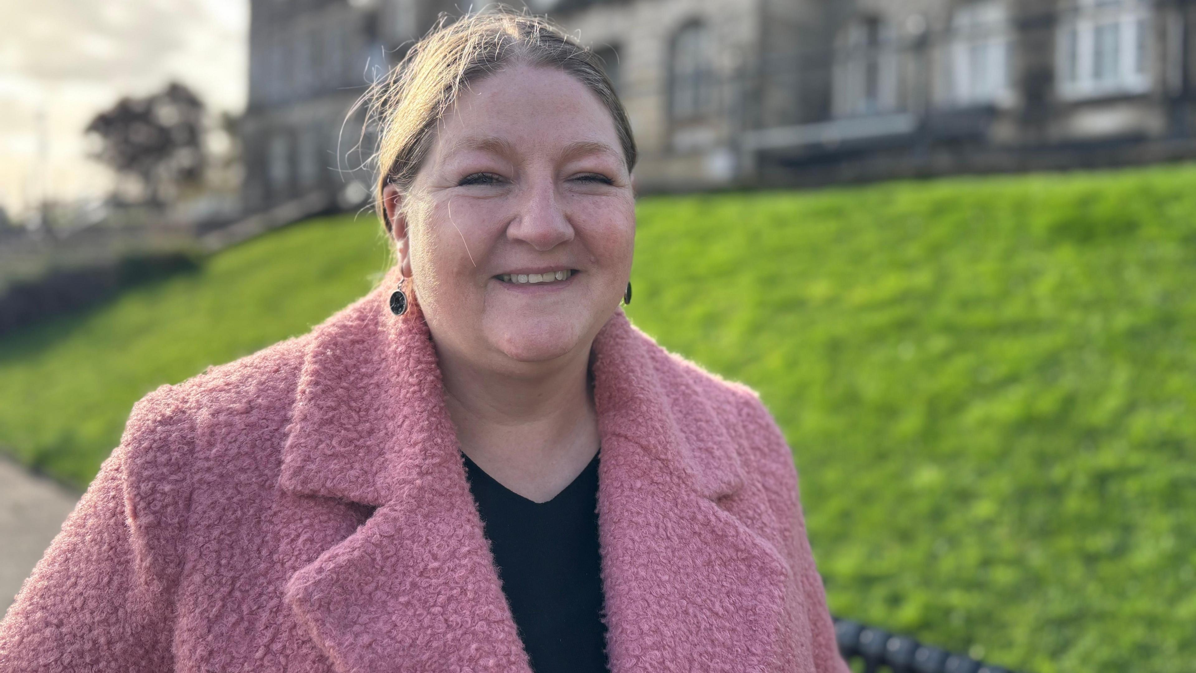 Councillor Ann Ainsworth smiles at the camera, her brown hair is pulled back from her face. She wears a pink coat and a black t-shirt.