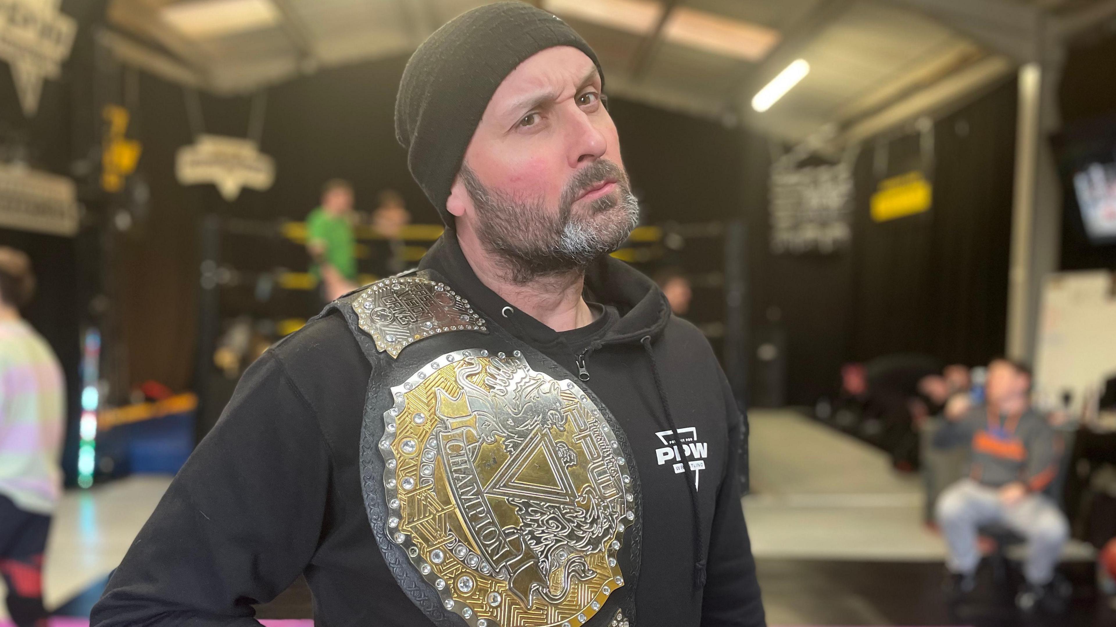 Man with beard wearing a black hat poses for the camera holding up a large gold wrestling belt