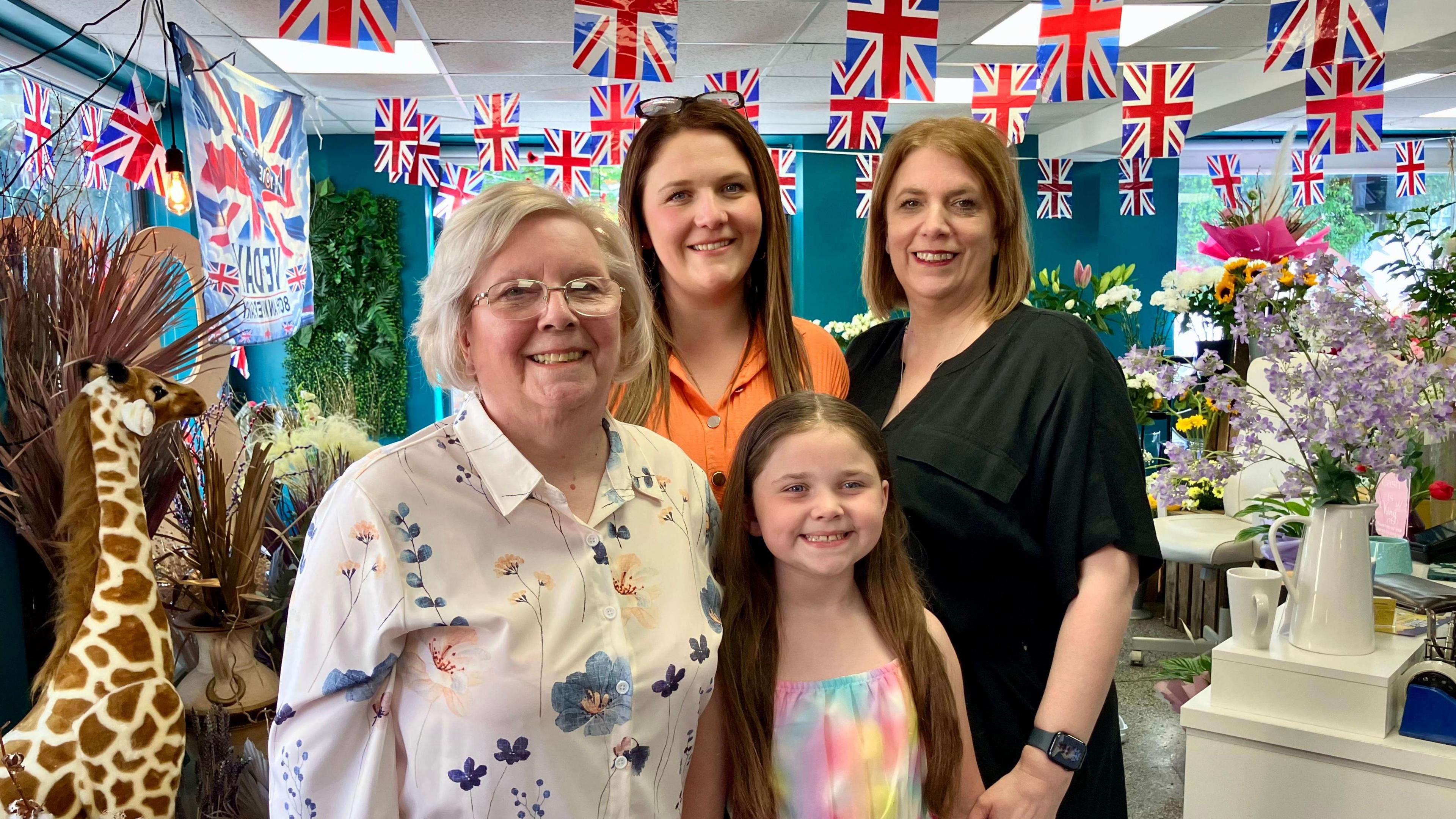 Three women of varying ages, and an eight-year-old girl, all from the same family, stand in a florist's shop. The shop is decorated for VE Day with union flag bunting.