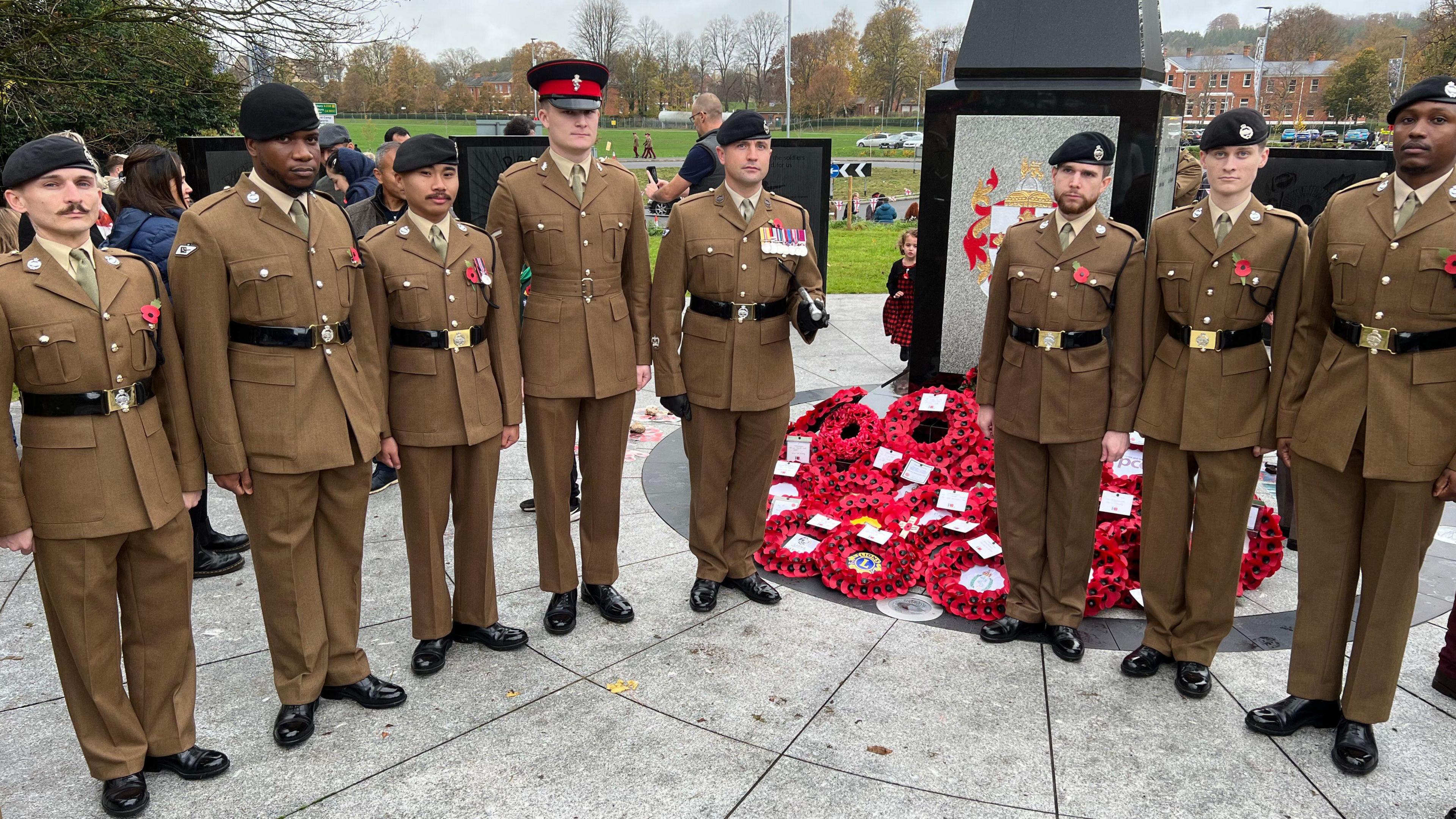 Eight men in front of a war memorial. They are all dressed in military uniform and looking straight at the camera with a blank expression.
