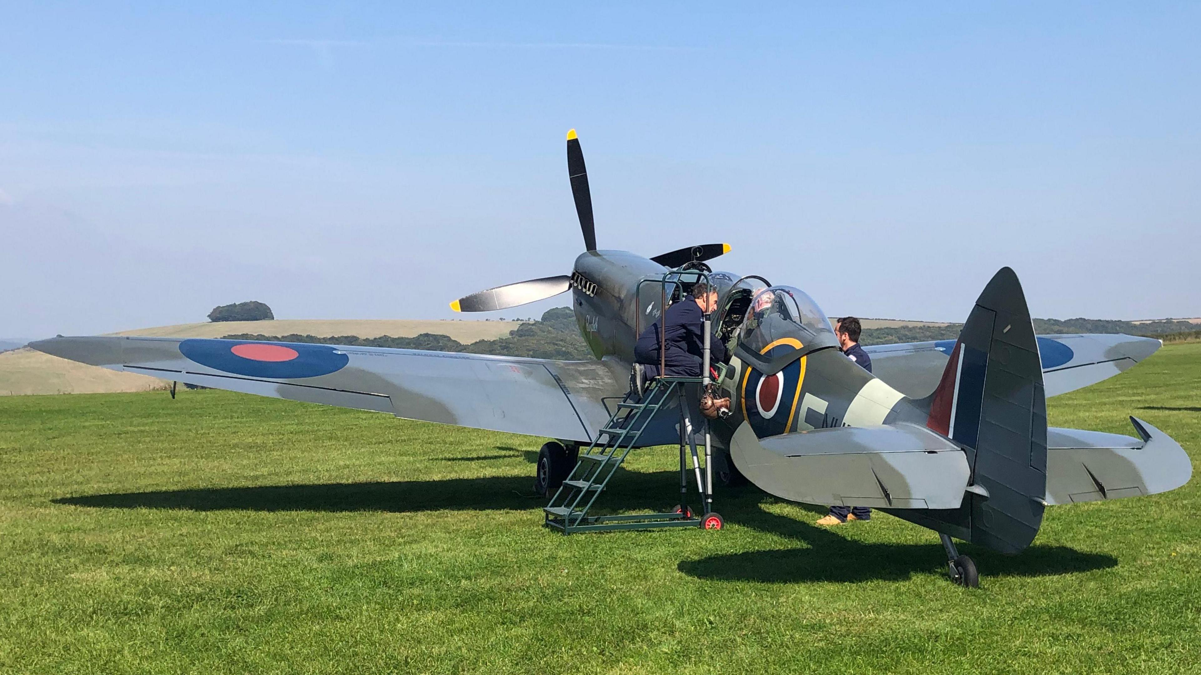 A spitfire on a grass field. Mr Fox is being fastened in his seat and briefed by officials.