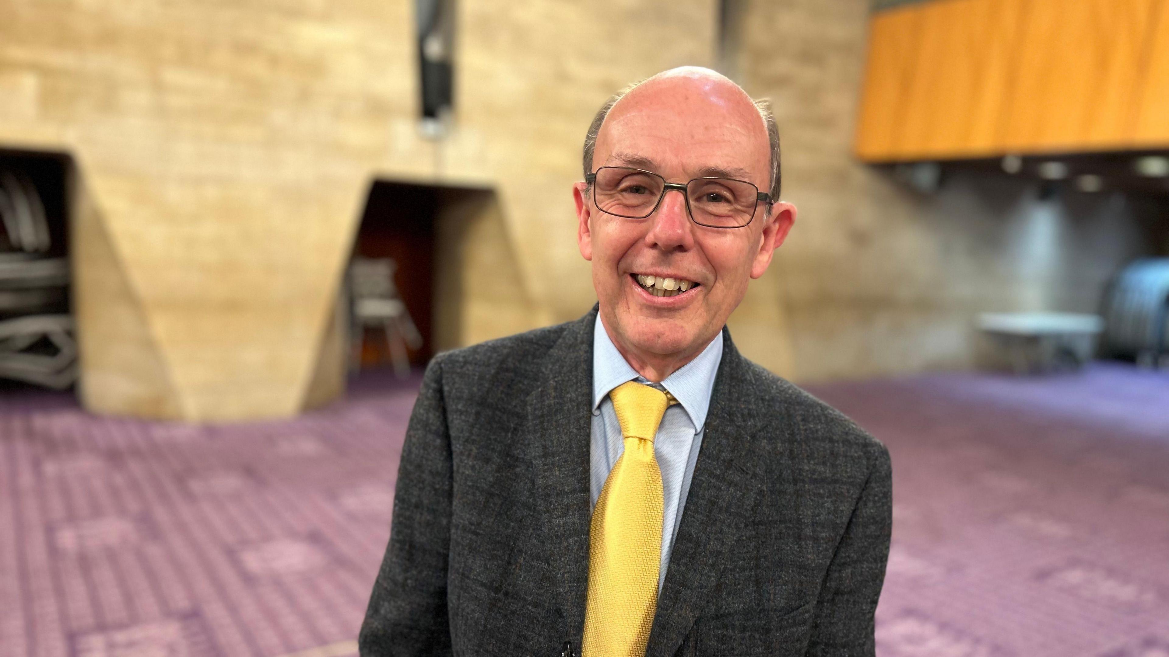 Man with glasses and suit and yellow tie looking at the camera.