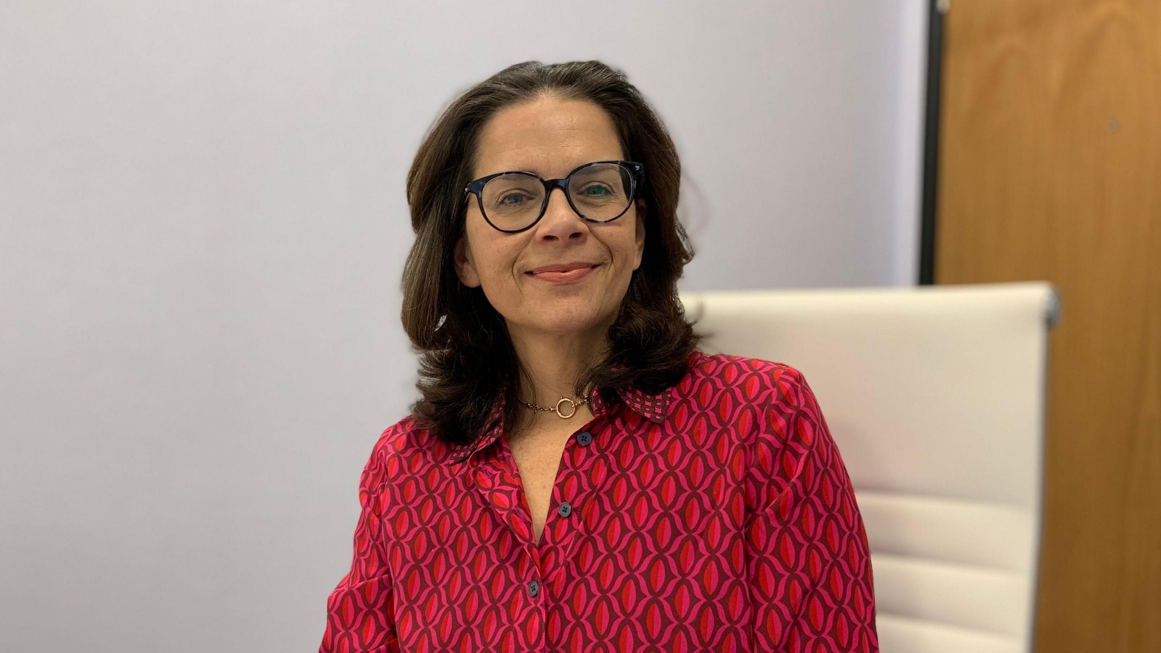 Deputy Dudley-Owen sat on a white chair. She is wearing a red patterned shirt. She had shoulder length brown hair, blue framed glasses and a gold necklace. 