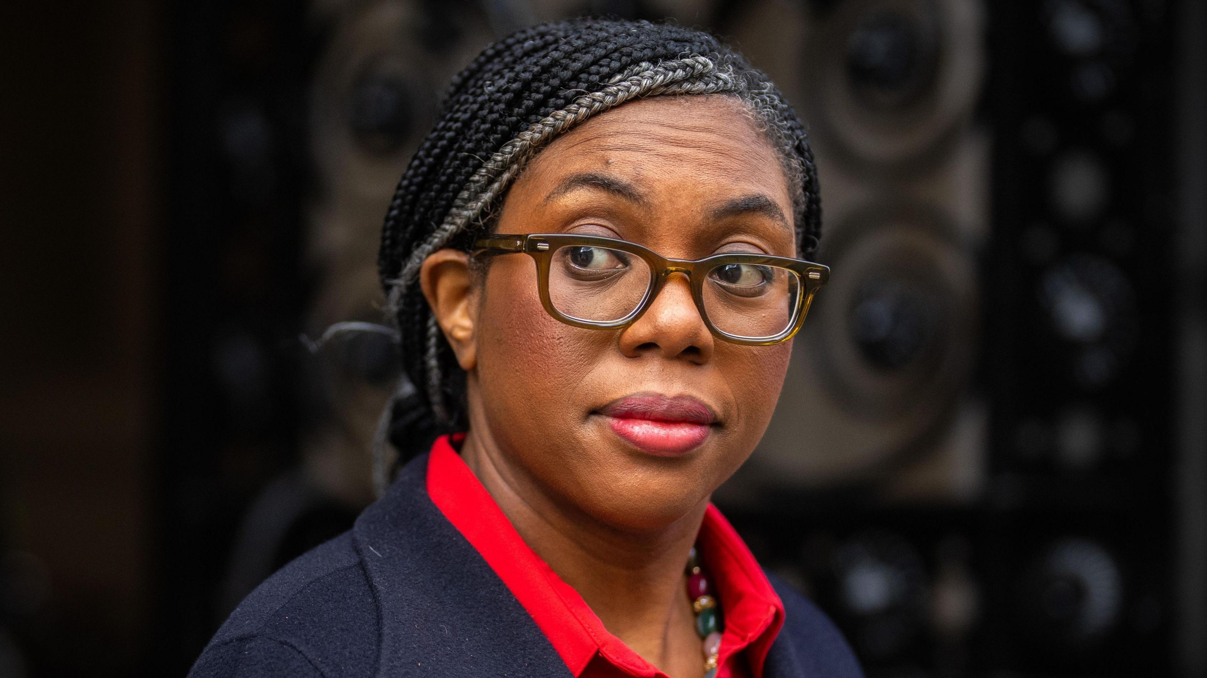 Kemi Badenoch wearing glasses and a red blouse looks at the media via the corner of her eye in Downing Street