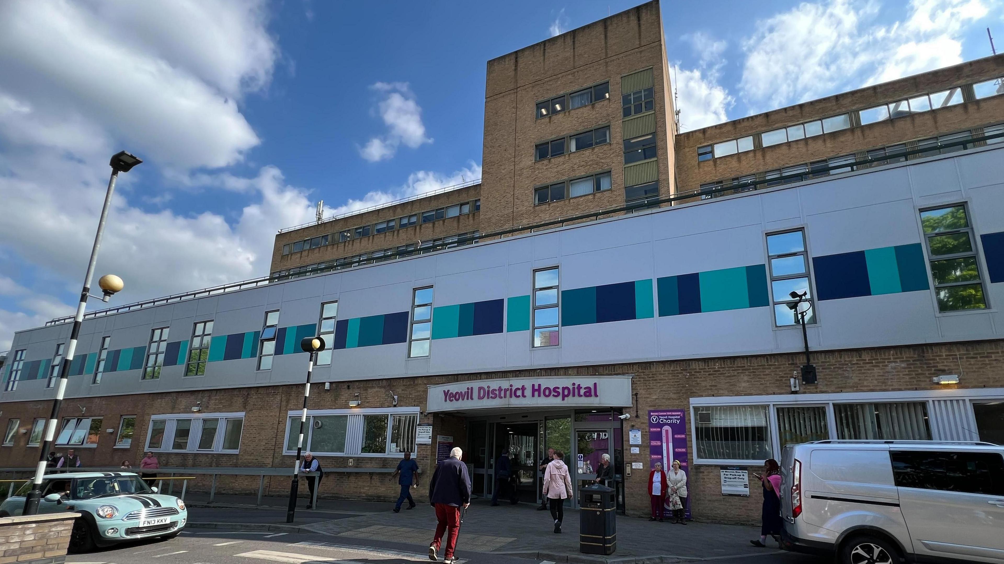 The picture shows the entrance to Yeovil District Hospital.  There are people walking on a crossing.  There are two vehicles outside the hospital.