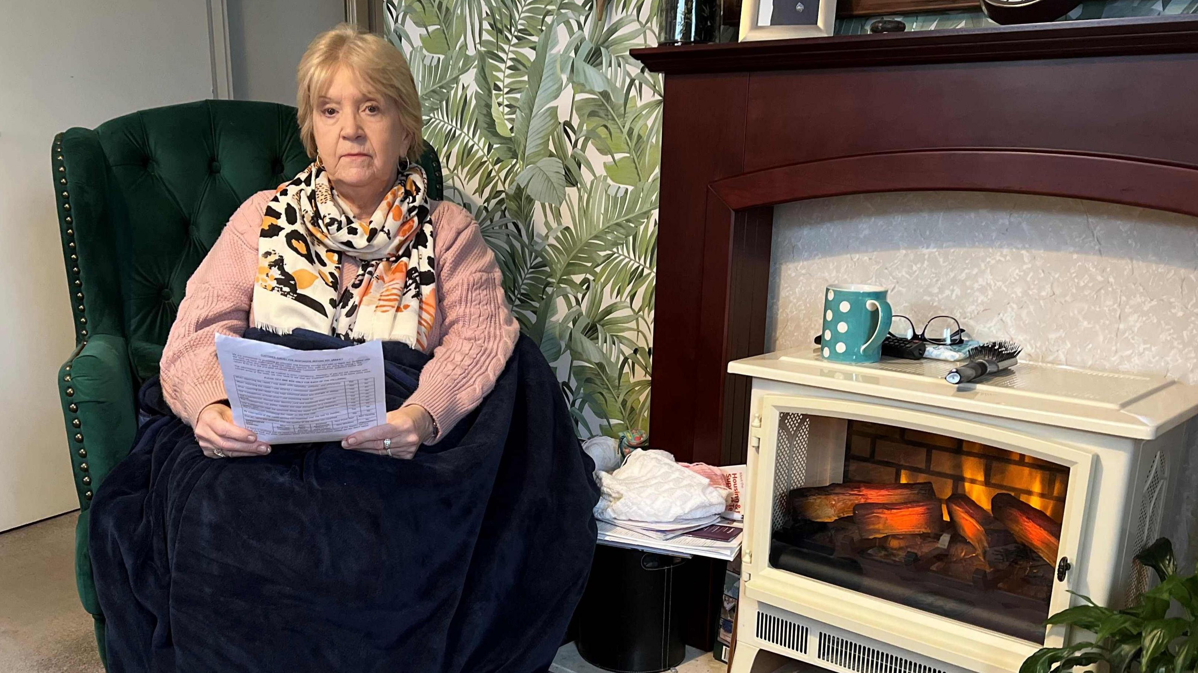 Tina Williams sitting in a green armchair, wearing a pink jumper, cream and orange scarf, a big, dark blue blanket on her lap and holding a letter from the council. 