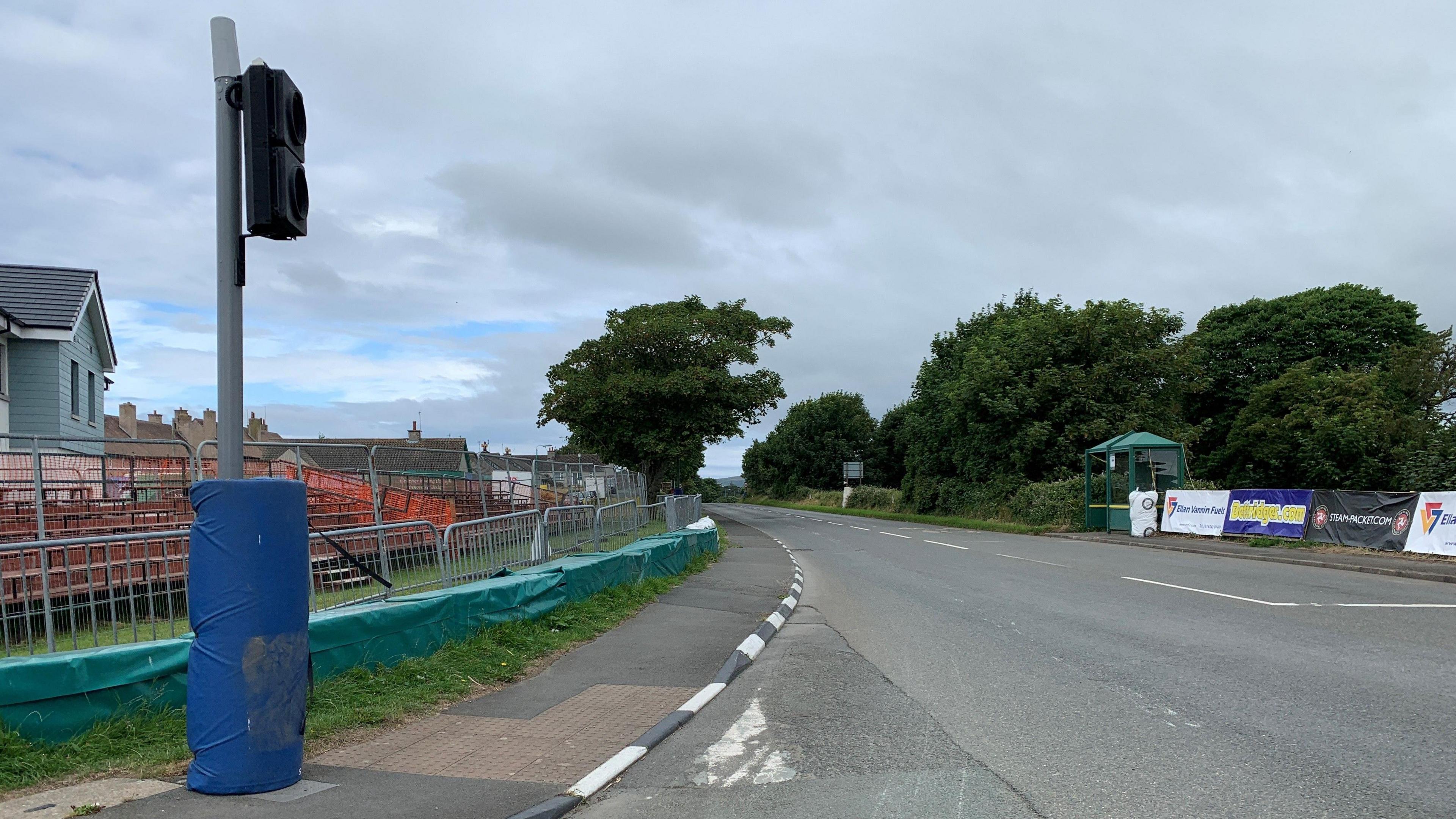 The road at Castletown Corner on the Southern 100 course. There are banners on the right and a small orange grandstand on the left behind fencing and barriers. There is a blue padding around the bottom of a traffic light pole.