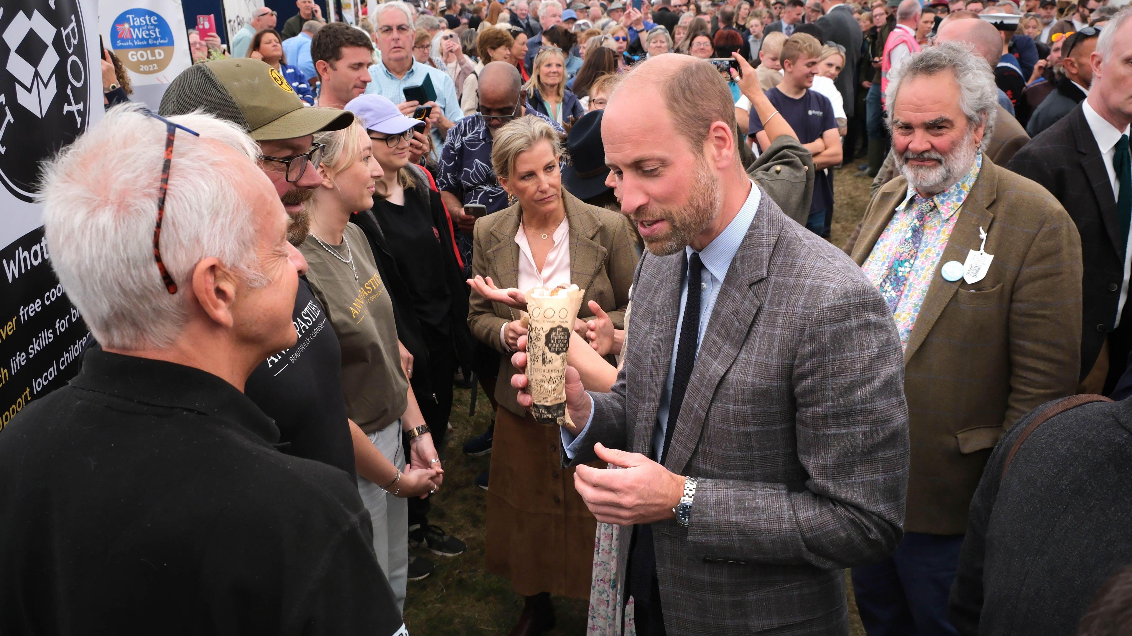 Prince William holds a pasty in his right hand while talking to two men at the Royal Cornwall Show. A large crowd has gathered around him and the Duchess of Edinburgh.