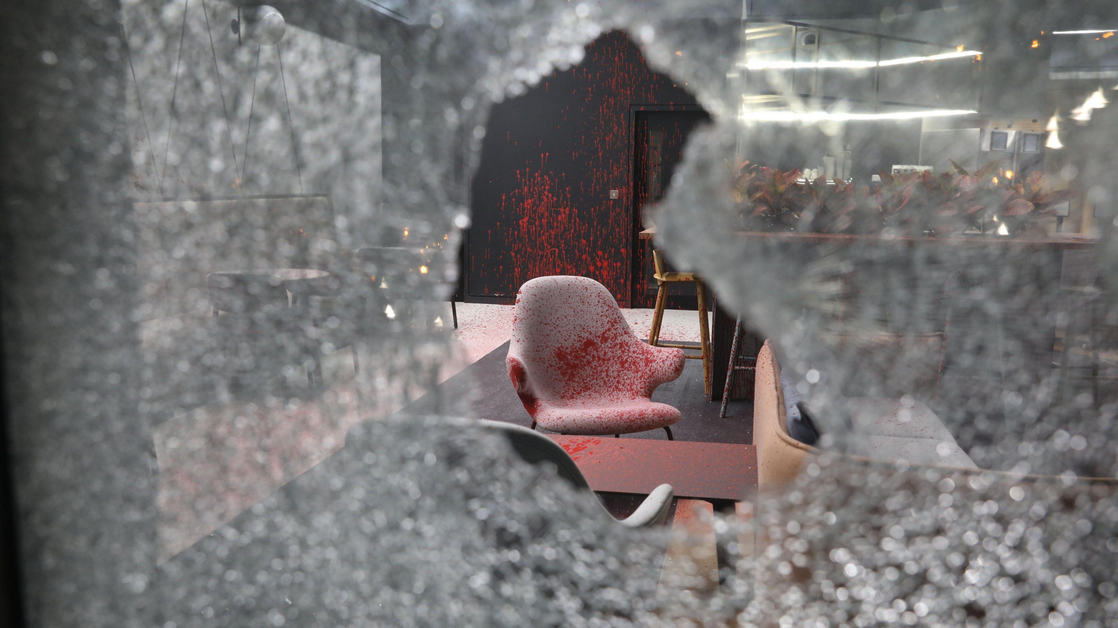 A photo of the red-sprayed office taken through a hole smashed in the glass window. A chair and coffee table are covered in red paint. A wall behind the chair is also sprayed in the paint.