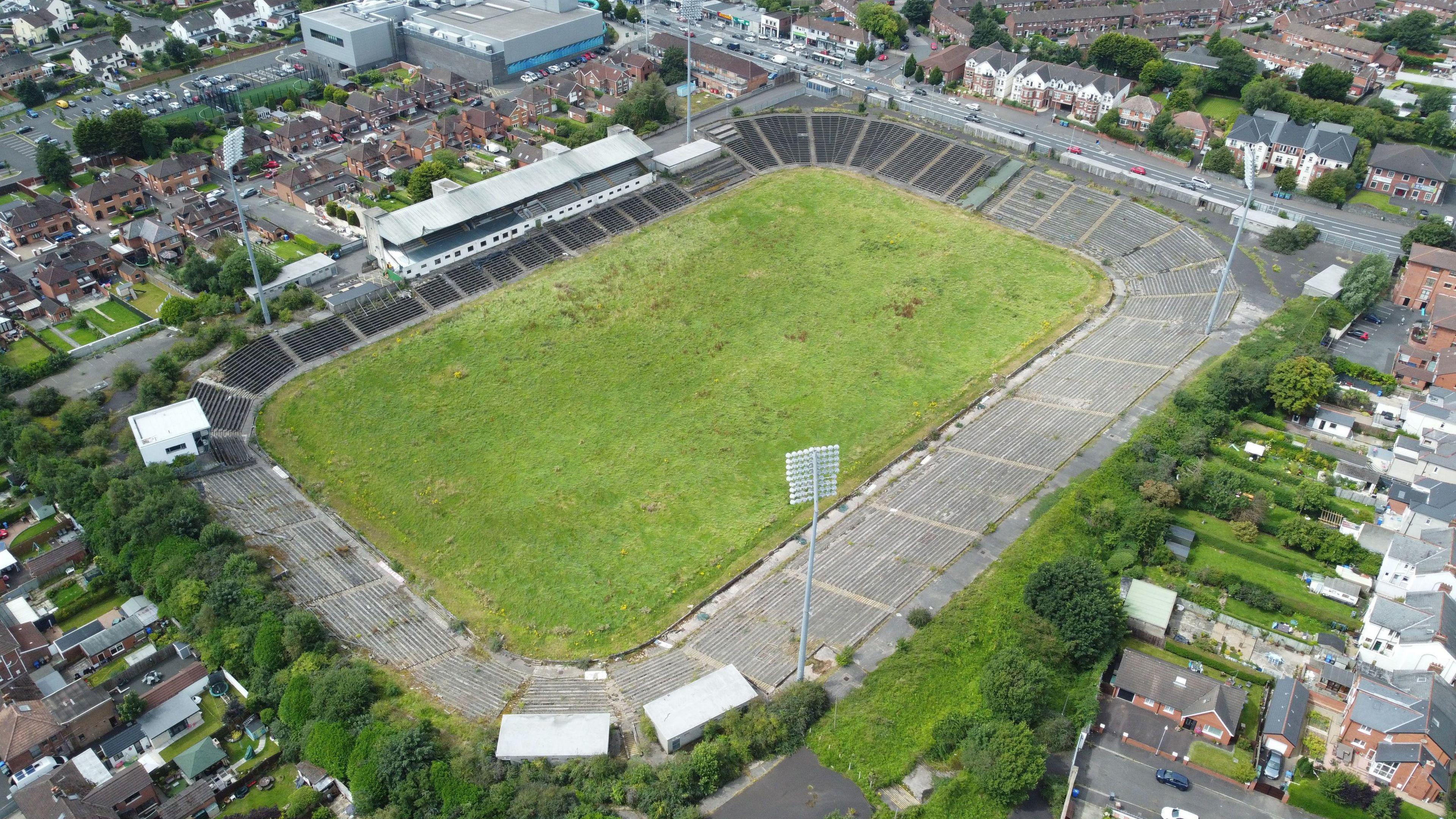 An aerial view of a stadium. The grass is in poor condition and looks overgrown, and the stands are dilapidated. Around the stadium are residential buildings and gardens.,
