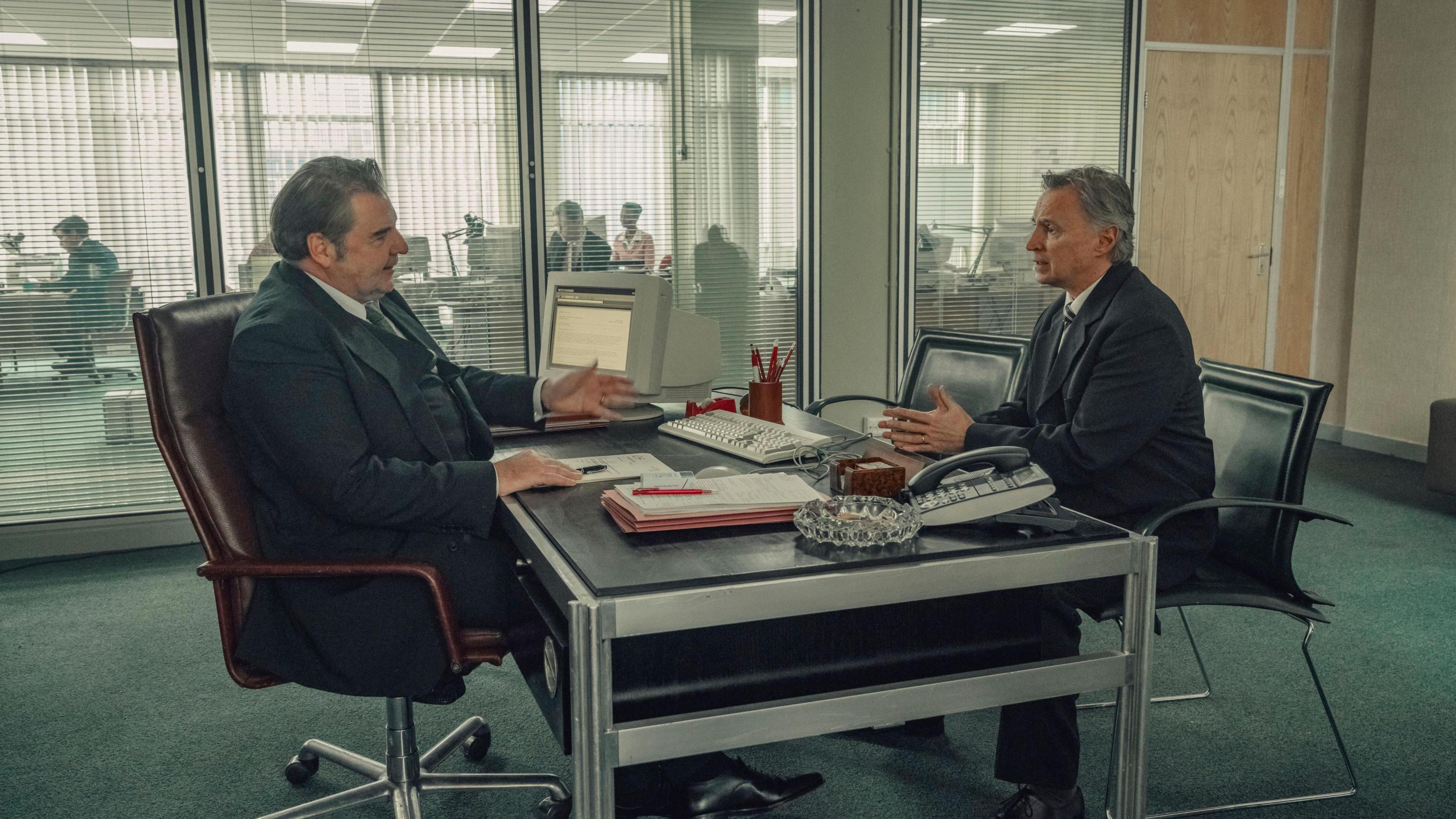 A shot from the Toxic Town television series. Two men wearing charcoal suits sit opposite each other across a black topped metal table. There is a computer, an ash tray, a computer keyboard and some paperwork on the table. 