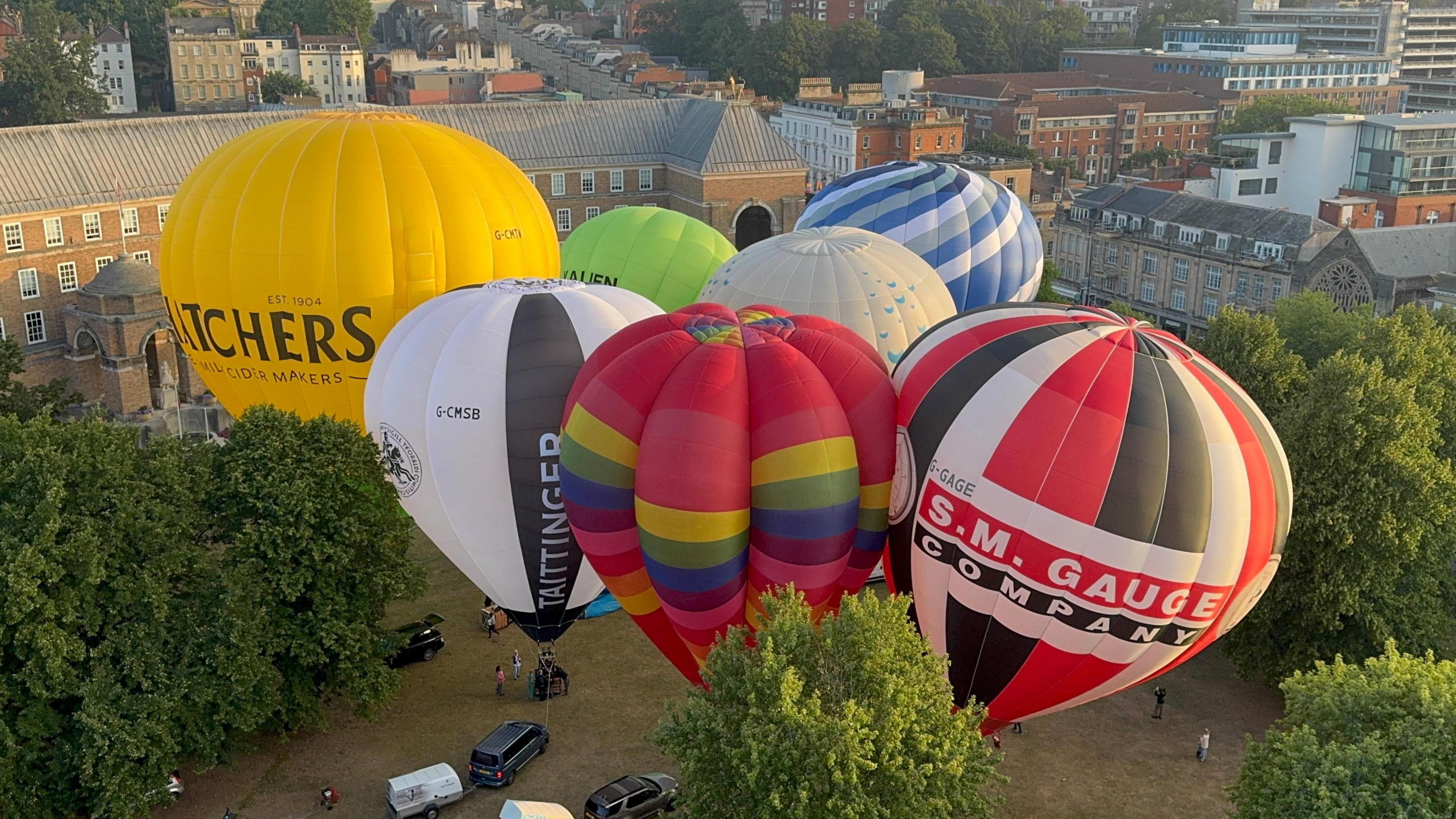 Several colourful hot air balloons inflated on the ground before taking off. The Bristol city landscape, including City Hall, can be seen in the distance behind them. There are also several vehicles parked near the balloons.