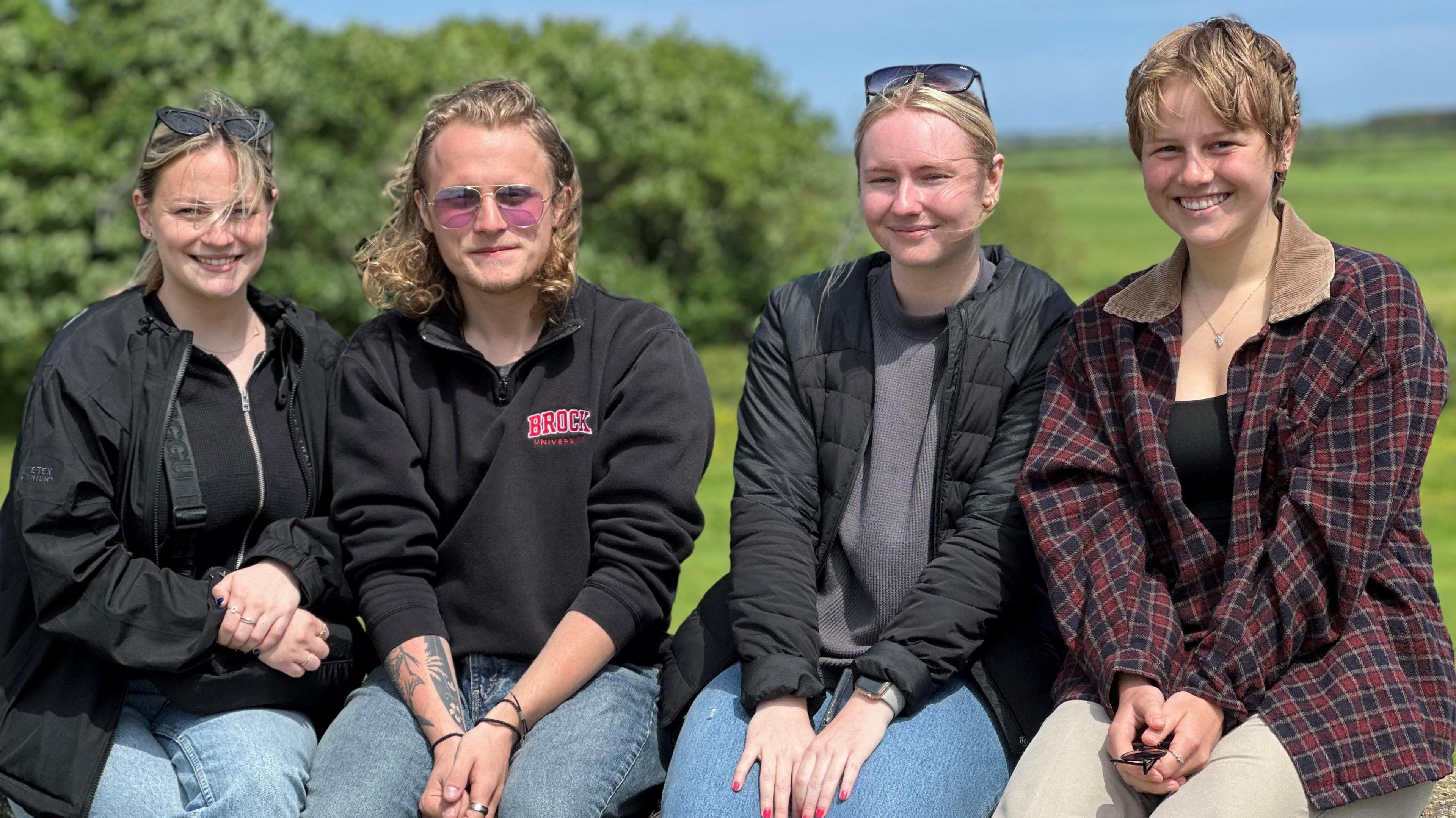 Four students sitting on a wall in a churchyard. There are green fields and bushes and a blue sky in the background.