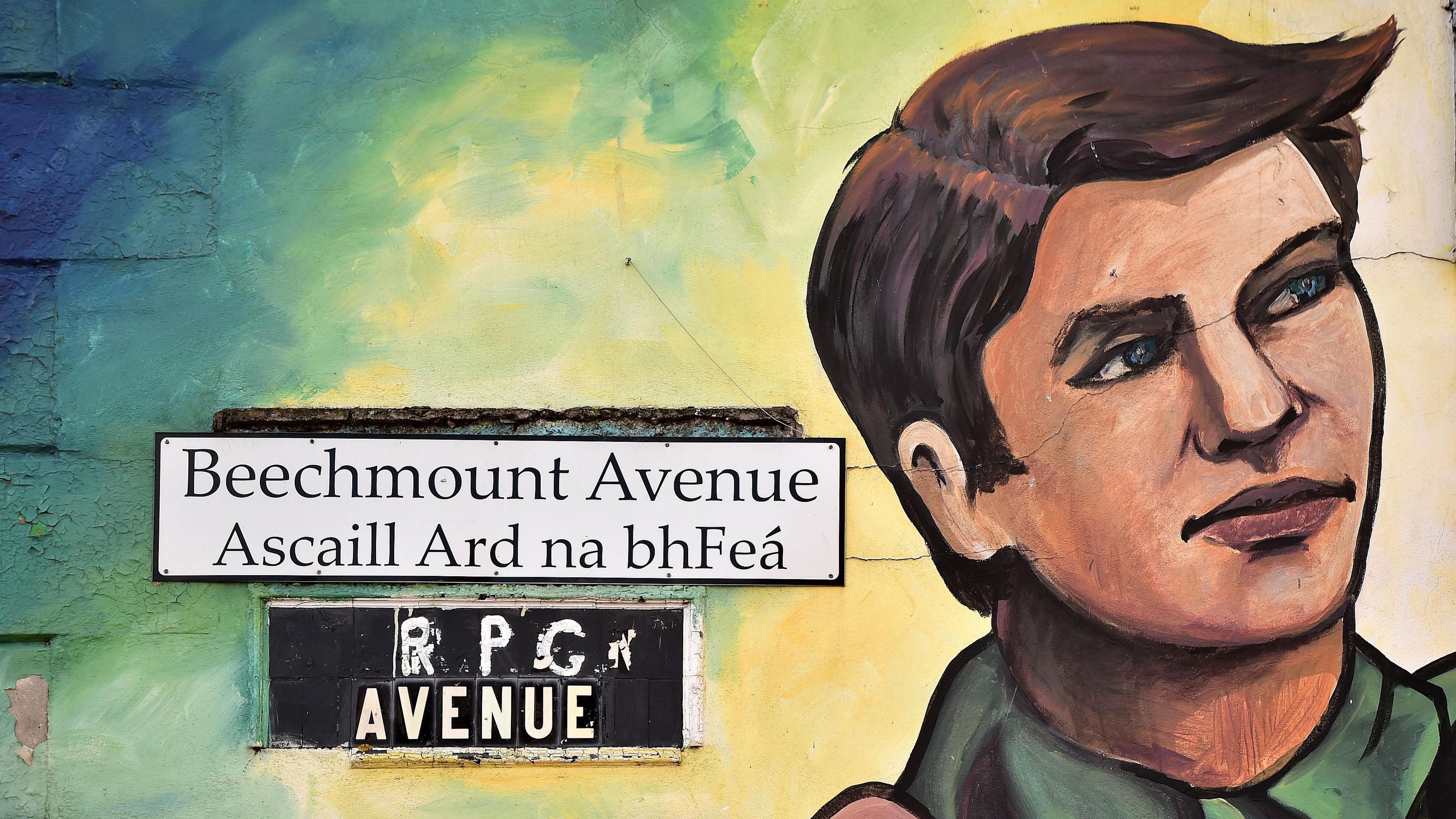 A dual English-Irish street sign for Beechmount Avenue is placed on a wall beside a painted mural of Michael Collins. Under Beechmount Avenue is "Ascaill Ard na bhFeá". 