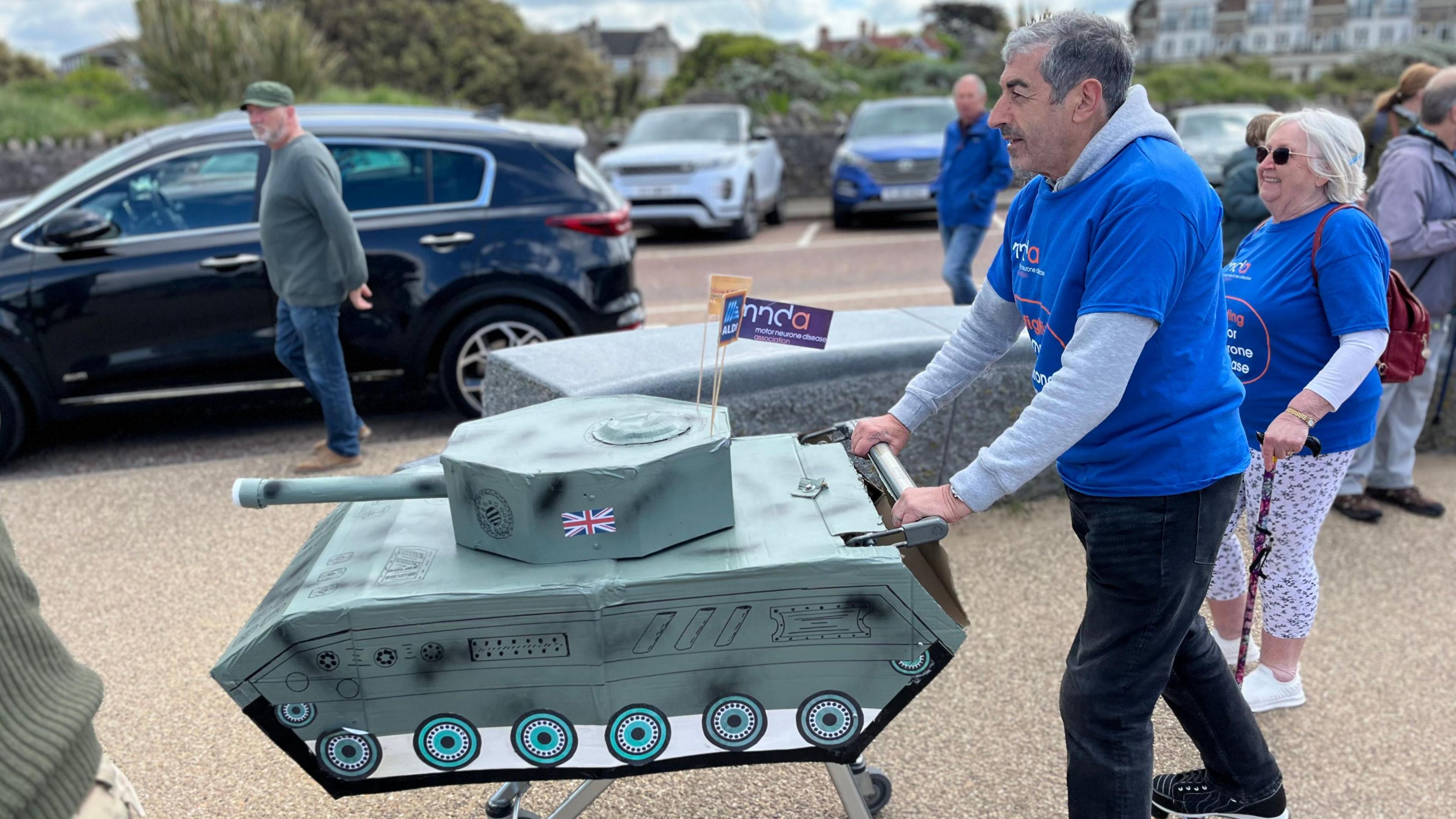 A man pushing a shopping trolley which has a cardboard tank mounted on it.