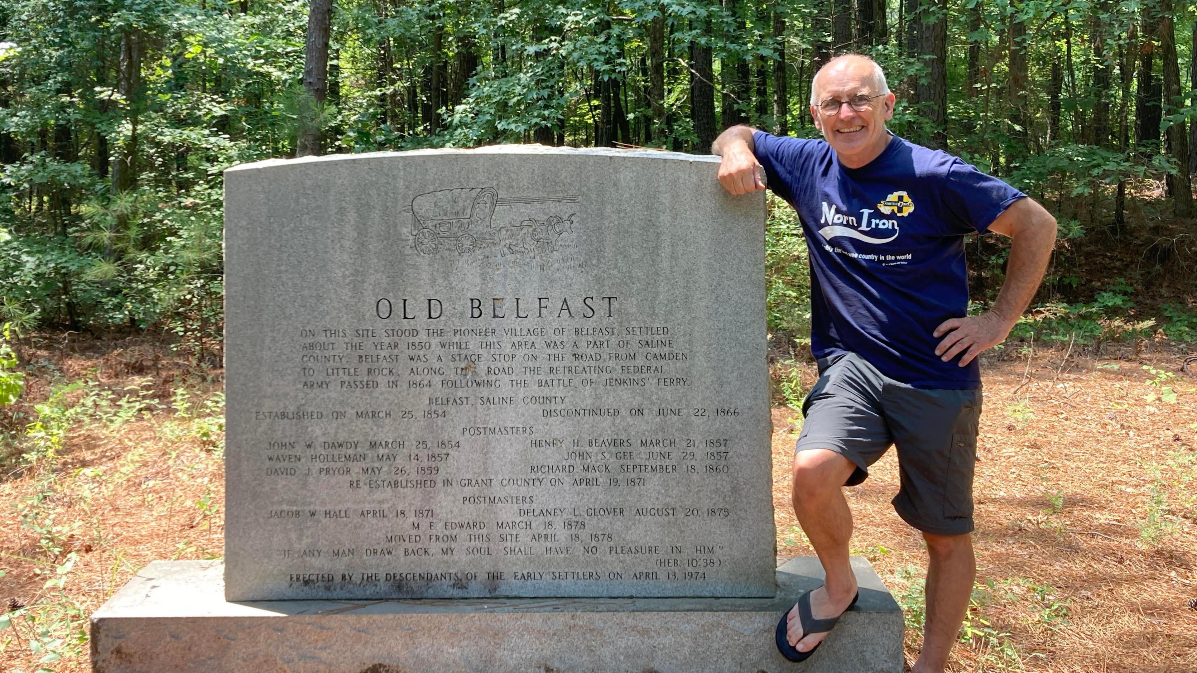 Sean Drysdale who is bald on top and has grey hair at the side and is wearing a Norn Iron blue T-shirt and grey shorts stands beside a a stone sign that says Old Belfast with a forest in the background