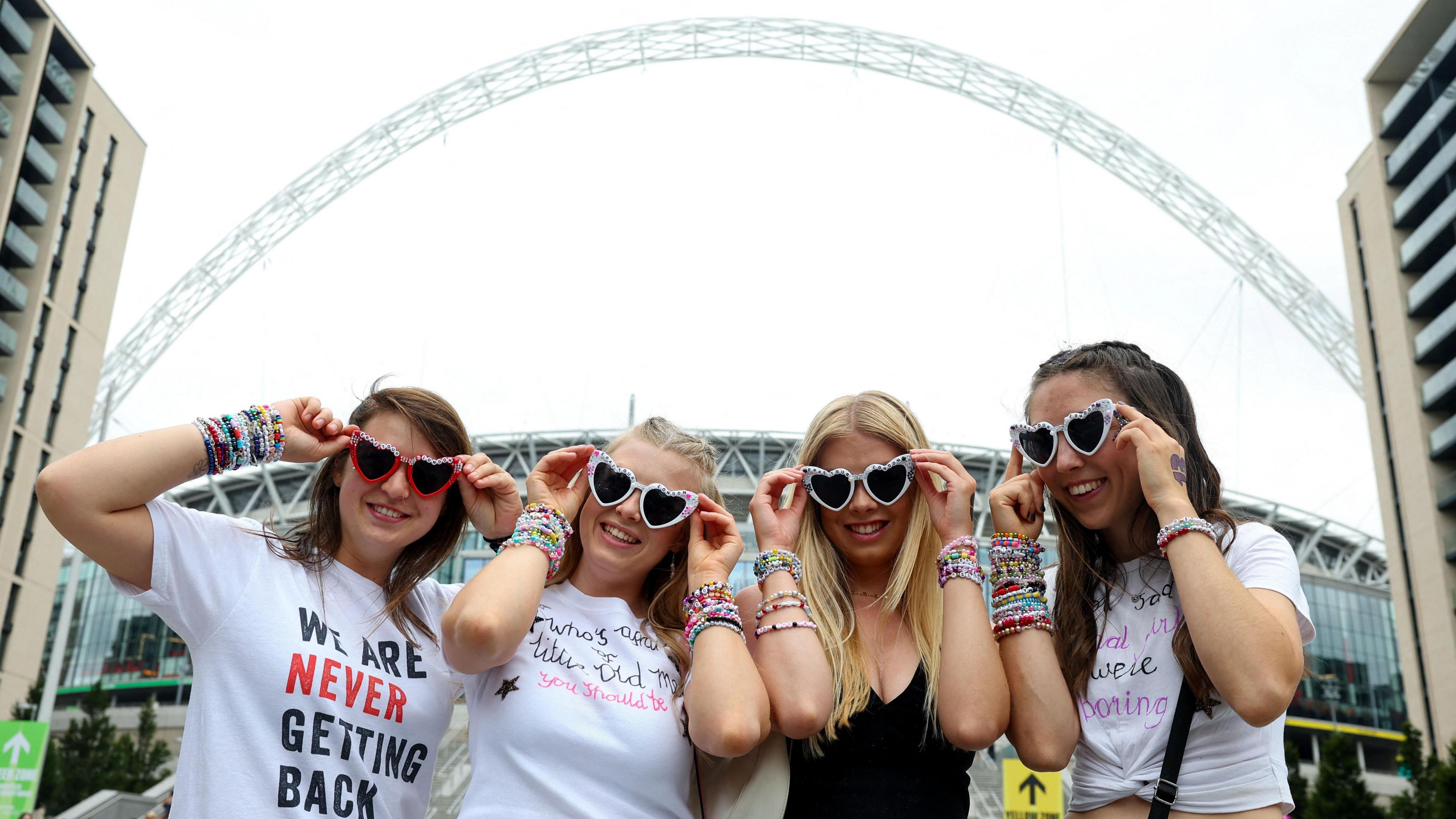 Infront of a backdrop of the arch at Wembley Stadium, four Taylor Swift fans post with their heart-shaped sunglasses and T shirts featuring song lyrics and phrases coined by the American singer. 