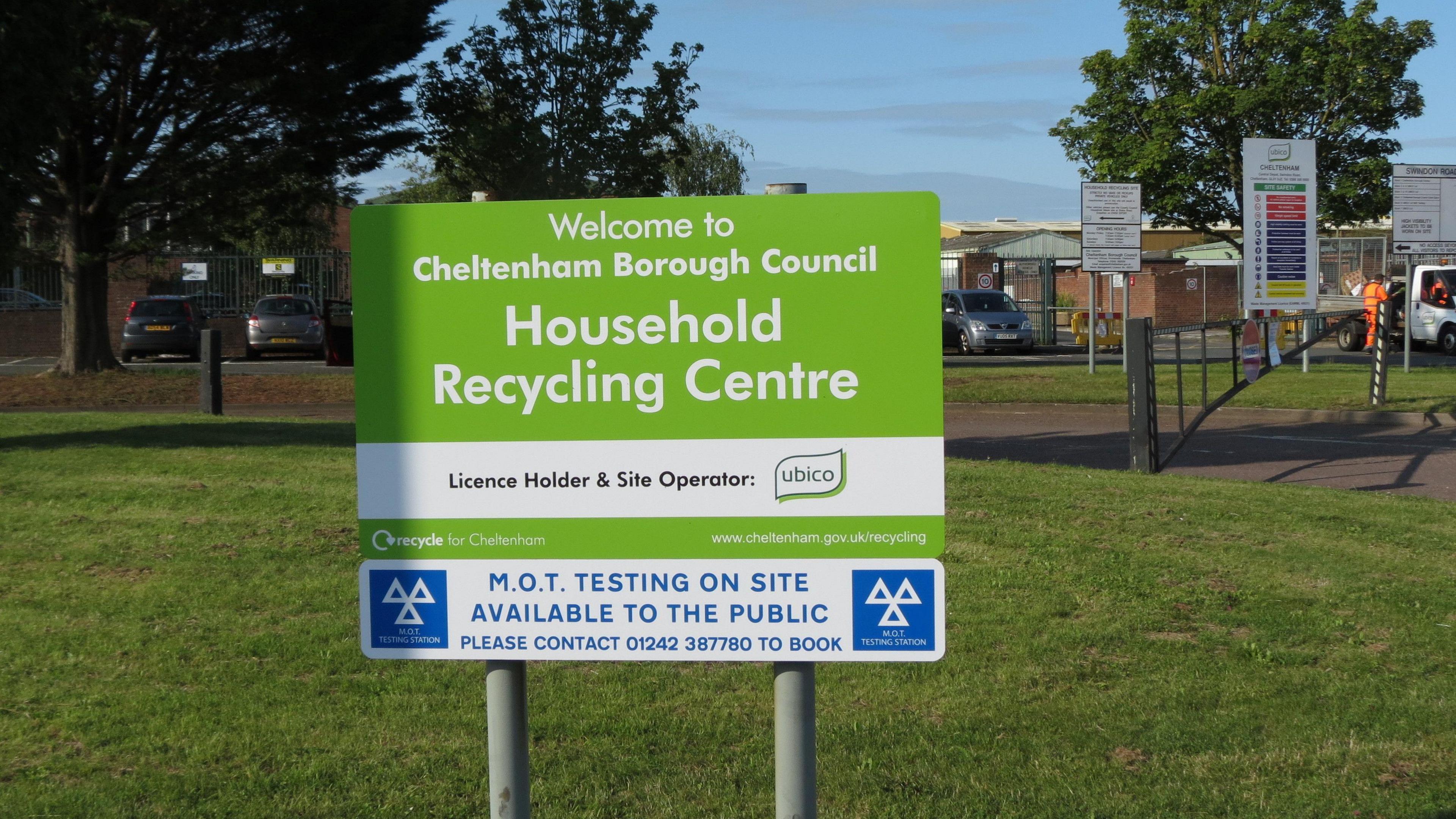 A green sign for Swindon Road Recycling Centre in Cheltenham that reads "Welcome to Cheltenham Borough Council Household Recycling Centre". The sign is on a grass verge at the front of the site. The recycling centre can be seen in the background along with a car park and men in orange hi-vis clothing in a van.