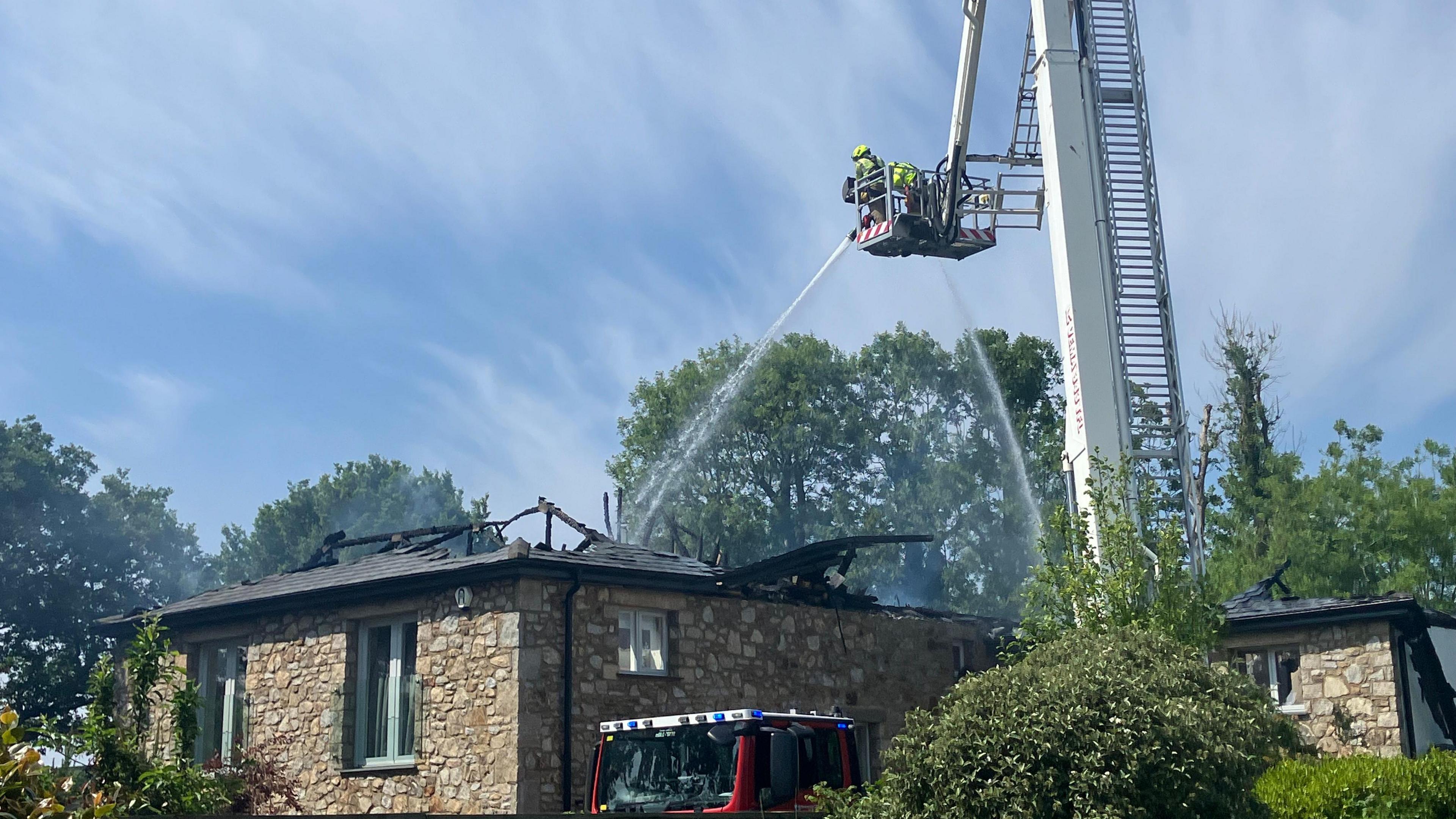 A ladder with two firefighters on it. They are spraying water on a house. No flames can be seen but there is smoke rising from the house.