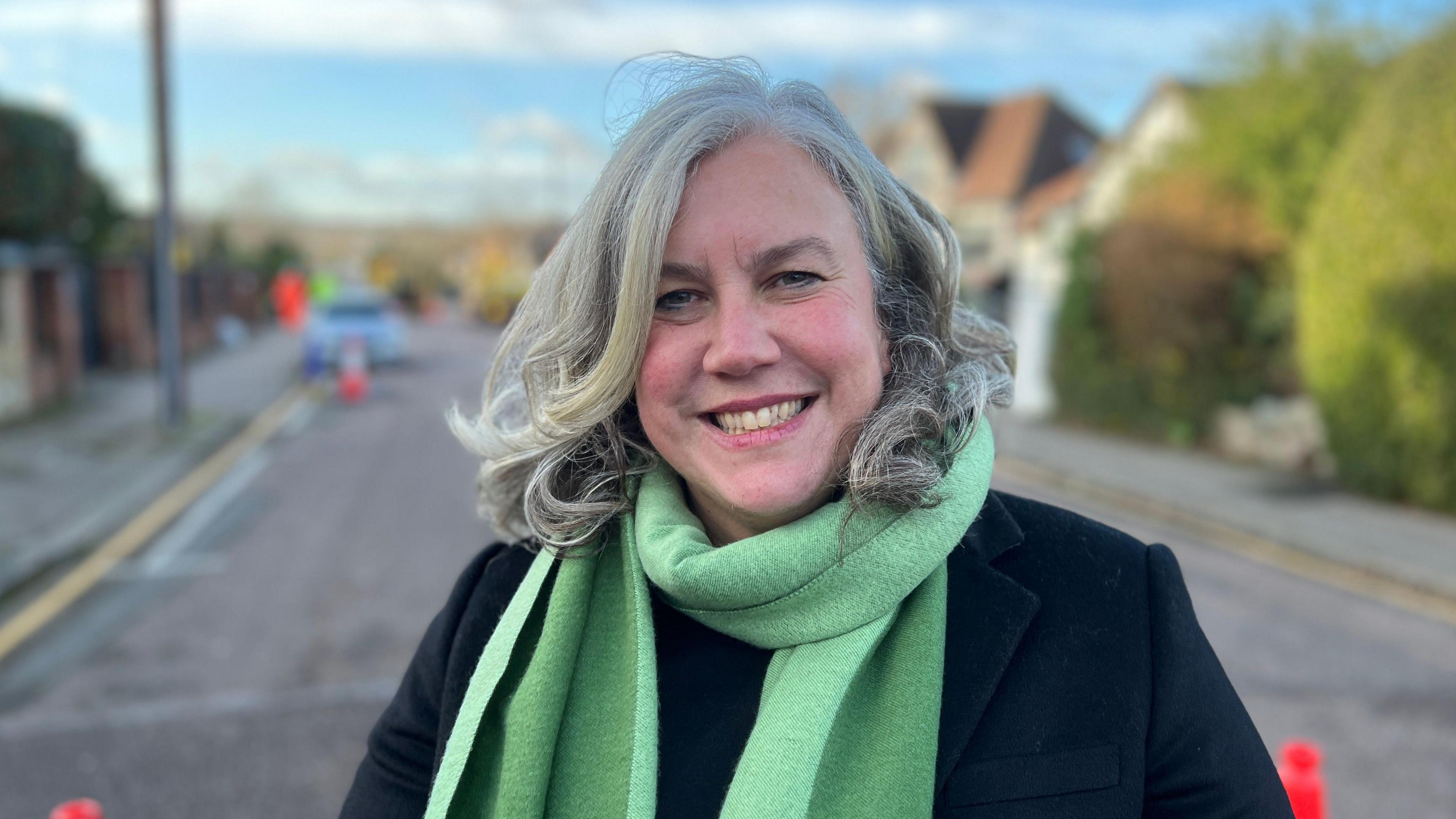 Heidi Alexander standing in a residential road. She is wearing a green scarf and navy blazer. There are red cones in the road. There are blue skies and white clouds.