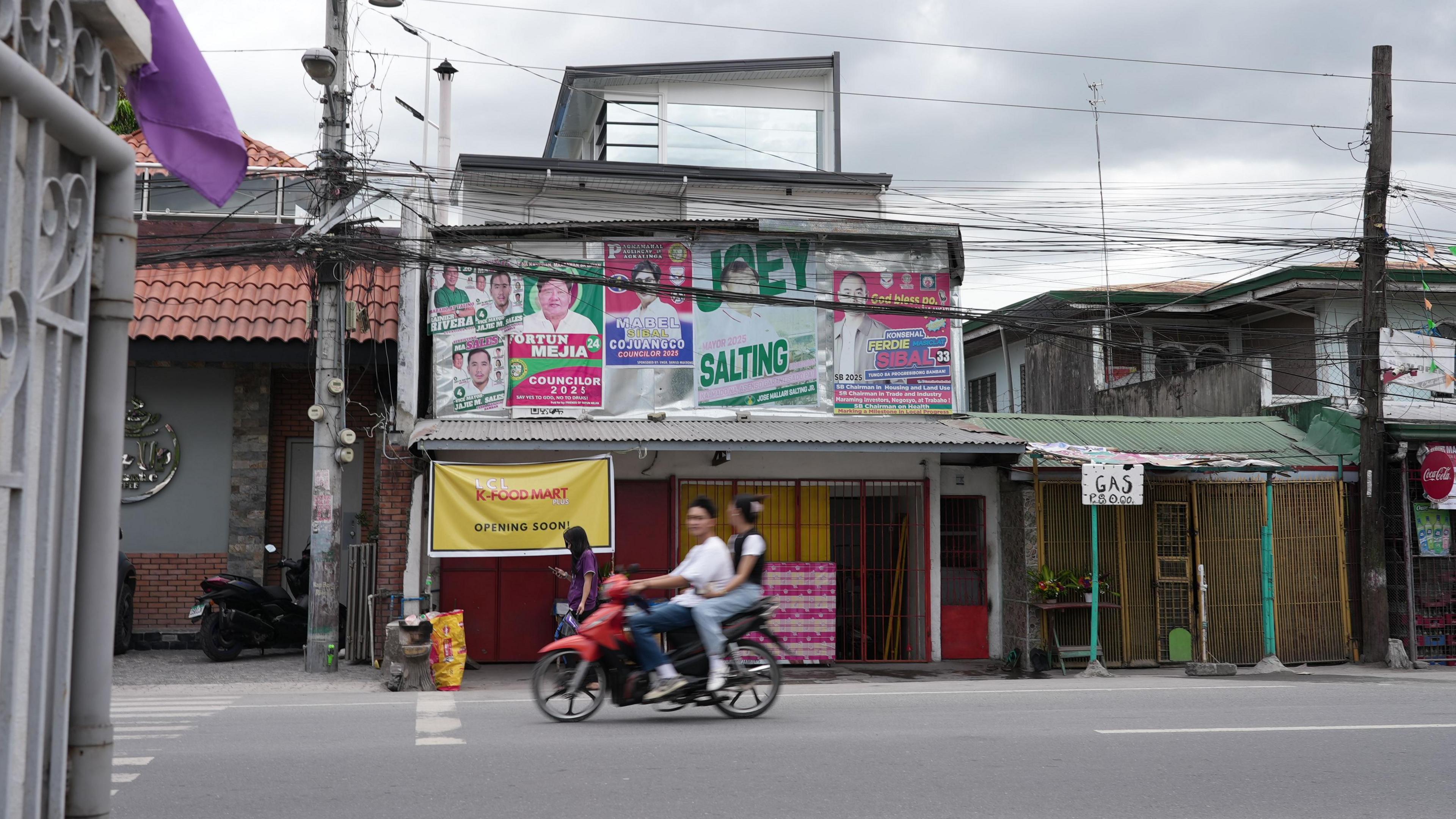 A couple on a moped ride past a shop plastered with green and pink campaign posters with the candidates' names and headshots