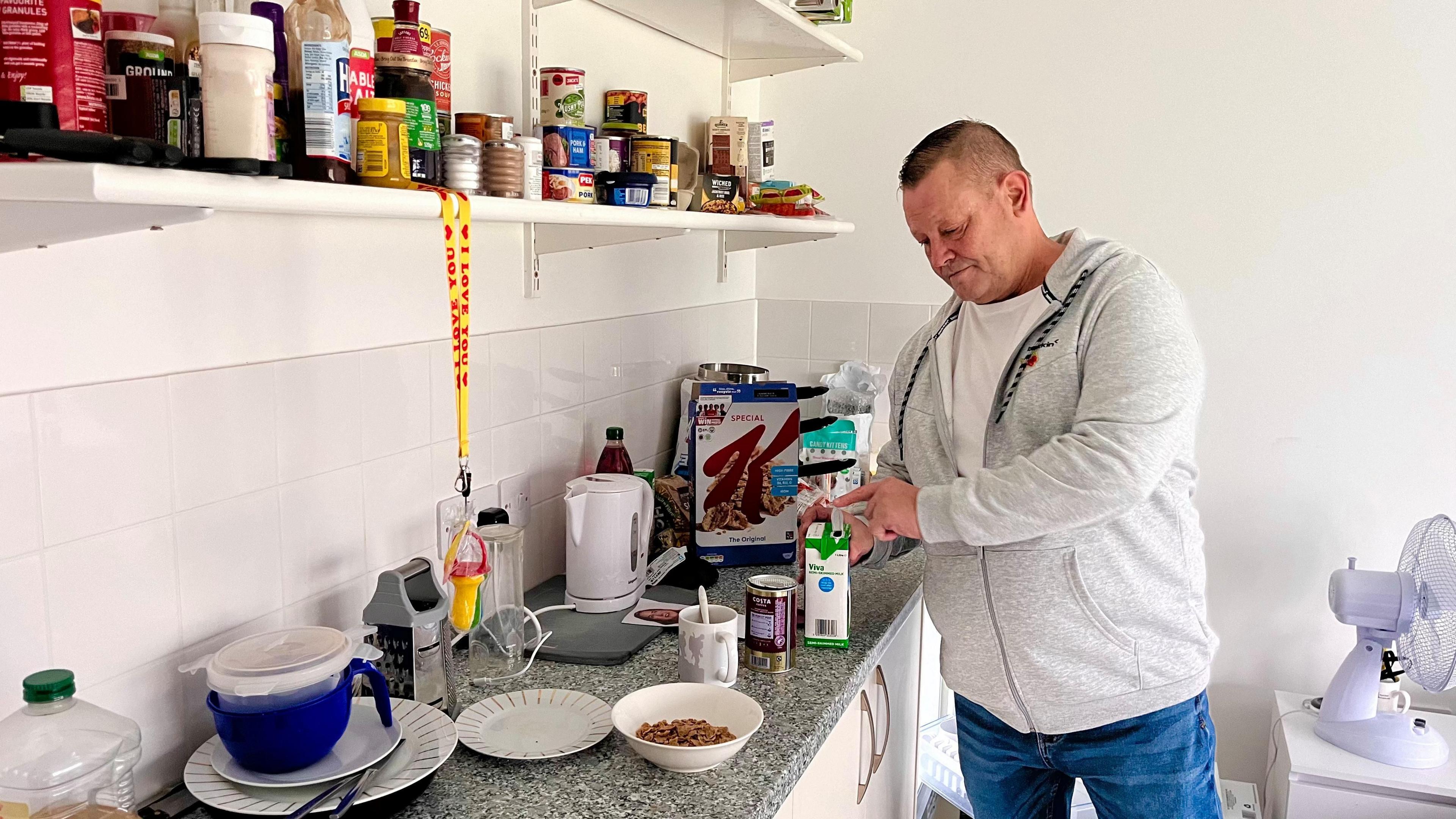 Gary James is wearing a grey hooded sweatshirt with a white T-shirt underneath. He is opening a carton of milk while standing in front of some kitchen units, about to make a bowl of cereal and a cup of tea