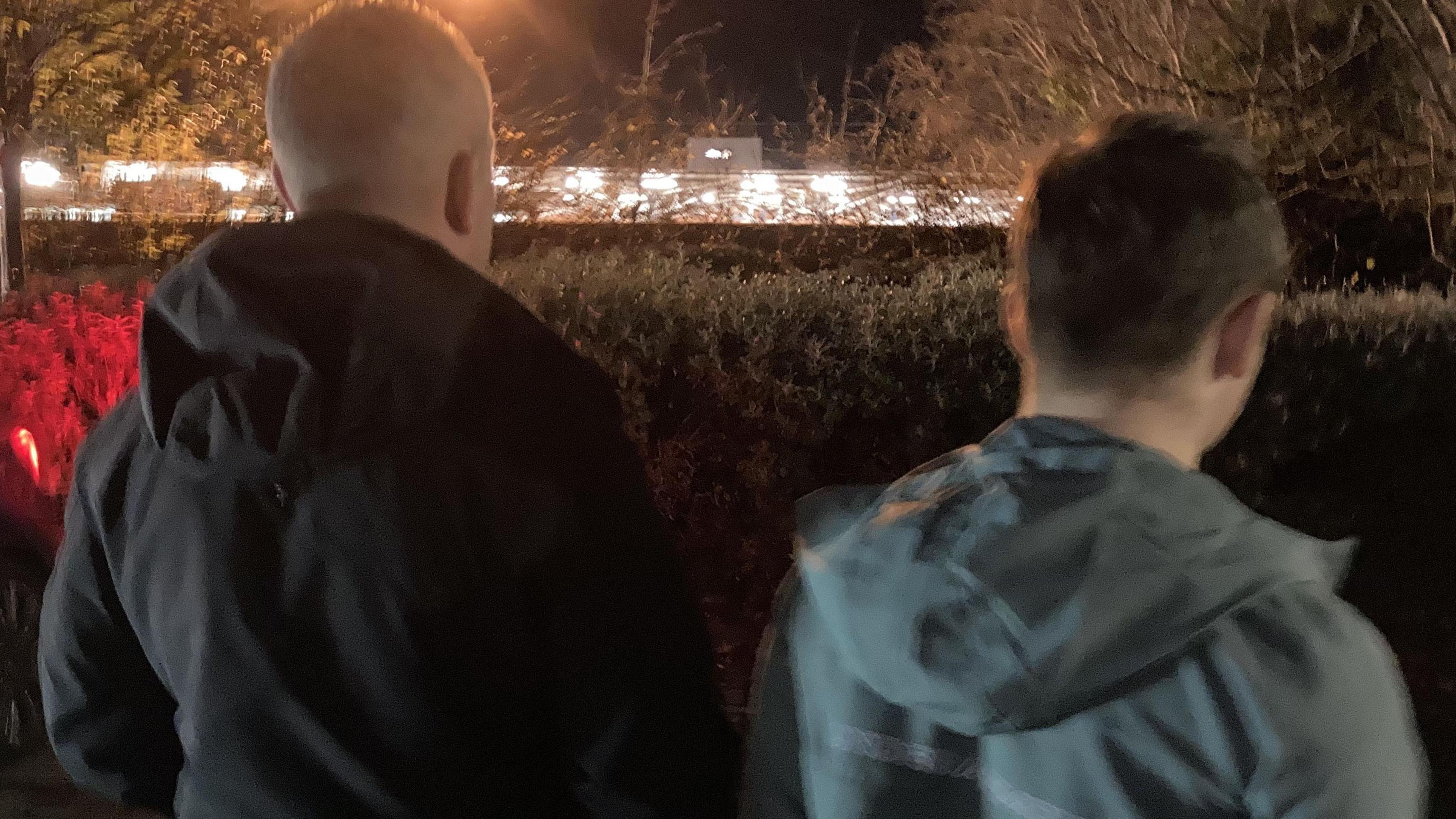 At night a man wearing a black jacket stands facing away from the camera, a teenage boy wearing a grey jacket stands next to him also facing away from the camera , a hedge is in front of them and lights from a car park illuminated by street lights is in the distance.
