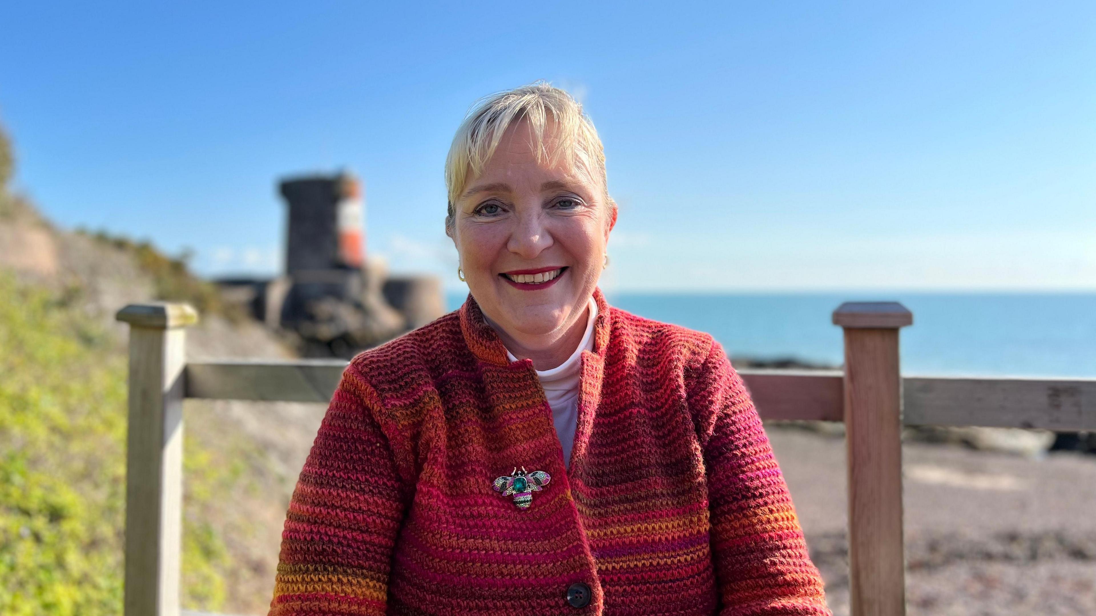 Tricia Warwick, a woman with blonde hair, wearing a red and orange top with a butterfly brooch on lapel. She is smiling, and sitting in front of a wooden fence, with the sea and beach visible behind her. To the left of the image, over her shoulder, is a Martello tower painted red and white.