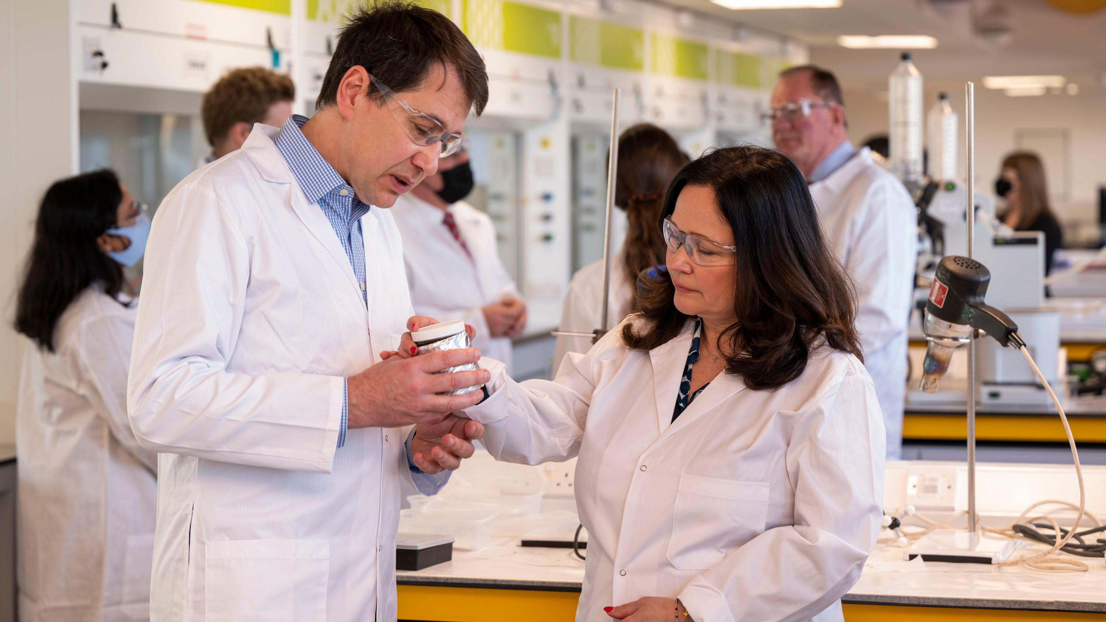 A dark haired man and woman in white lab coats and goggles in a research lab