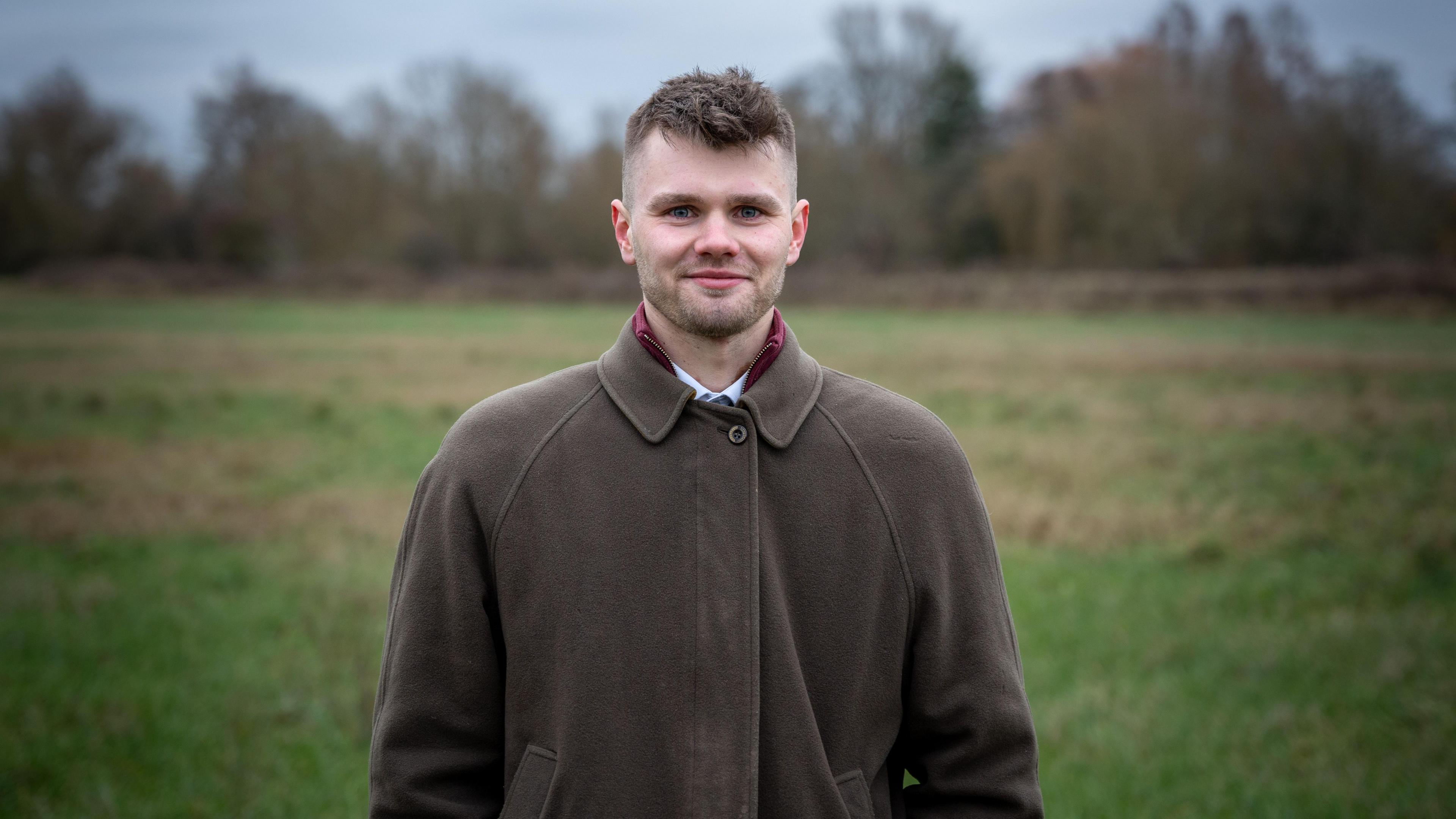 Dominic Mills standing in a field on a dreary day. The sky behind him is grey. He has short brown hair which is shaved on the sides, is wearing a brown jacket with a collared white shirt, and is smiling at the camera.